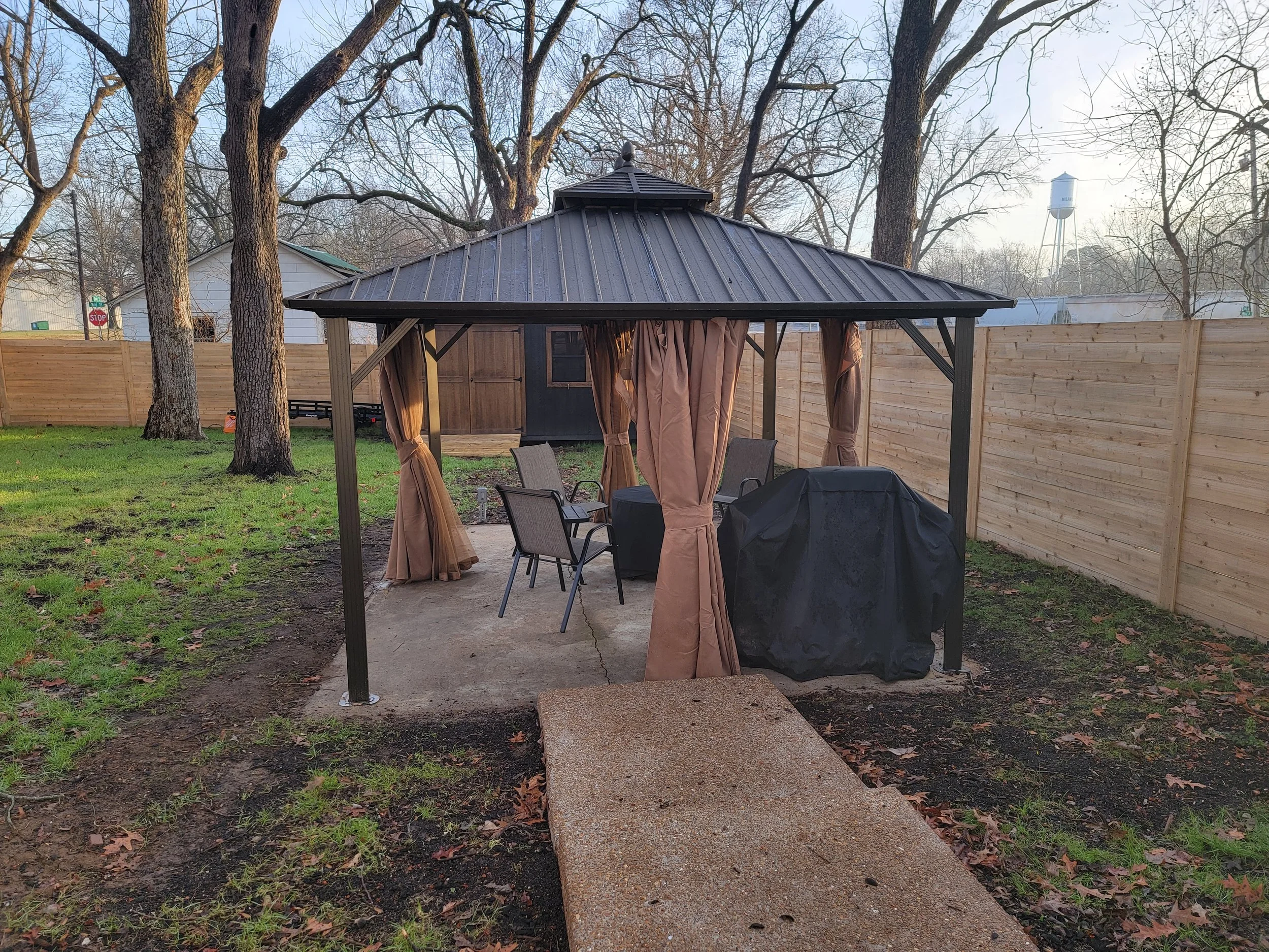 Backyard patio with a gazebo, outdoor table, and chairs, surrounded by a wooden fence, trees, and a shed in the background.