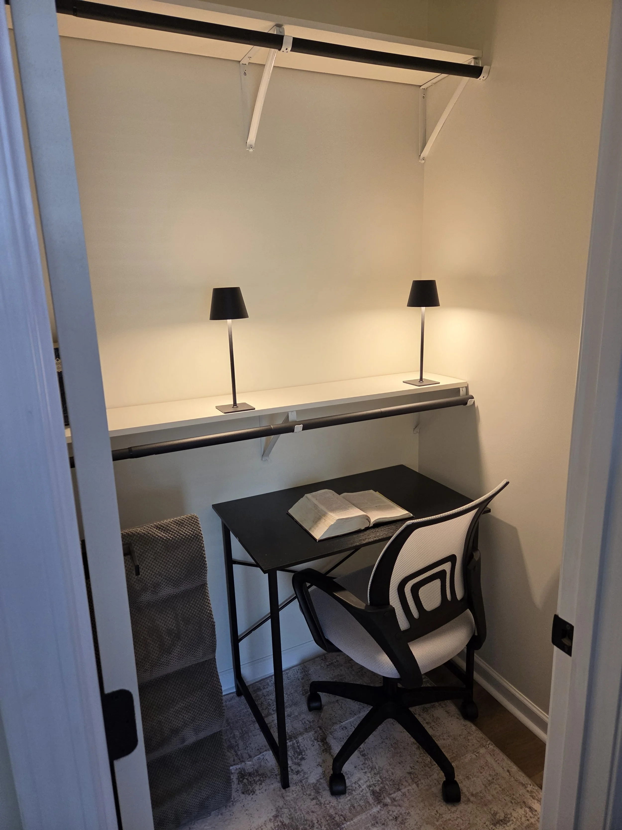 Small home office with a black desk, a white ergonomic chair, two small black lamps, an open book, a mesh-folding screen, and a built-in closet with hanging rods.
