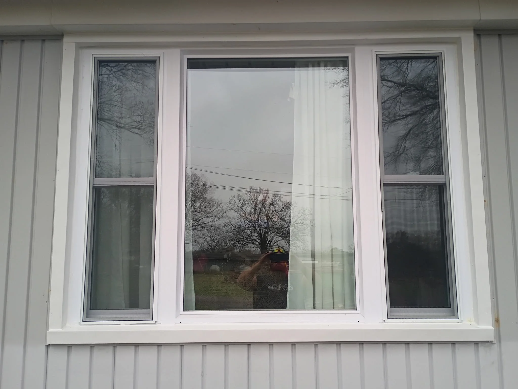A large rectangular window with four sashes on a white building exterior, reflecting trees and an overcast sky.
