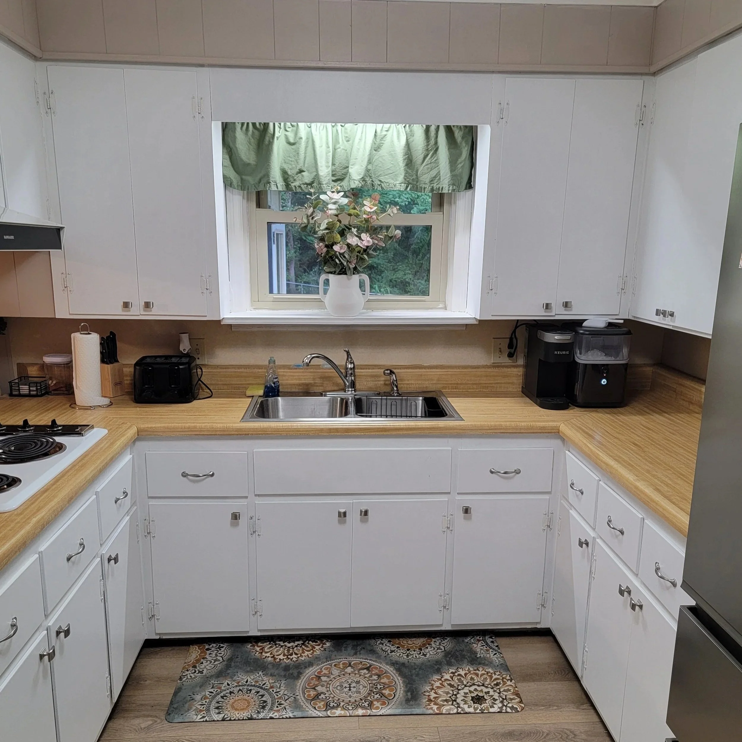 Kitchen with white cabinets, wooden countertops, a window with green curtain, and a vase with pink flowers on the windowsill. There is a stainless steel sink with a faucet, a small black box, a white paper towel roll, and a few small kitchen appliances on the countertops. A decorative rug is on the floor.