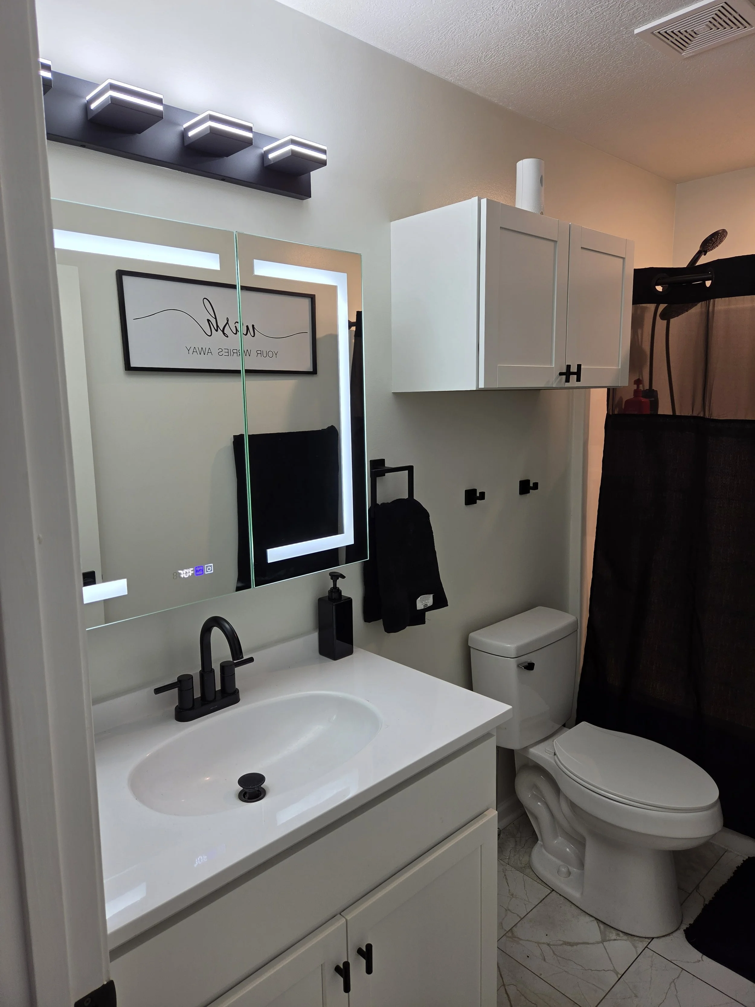 A compact modern bathroom featuring a white vanity with a sink, a mirror with LED lighting, a black faucet, black accessories, a wall-mounted cabinet, toilet, and a shower area with a black shower curtain and showerhead.