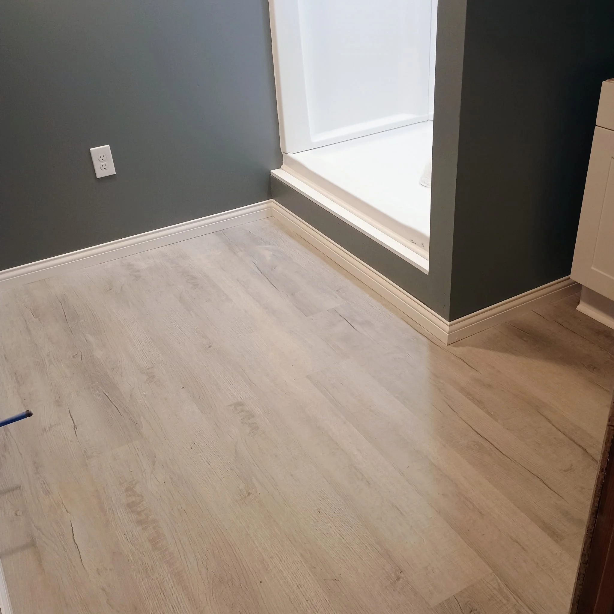 A corner of a room showing a gray wall with a white electrical outlet, white baseboard moldings, a section of a white shower stall, and light-colored wooden flooring.
