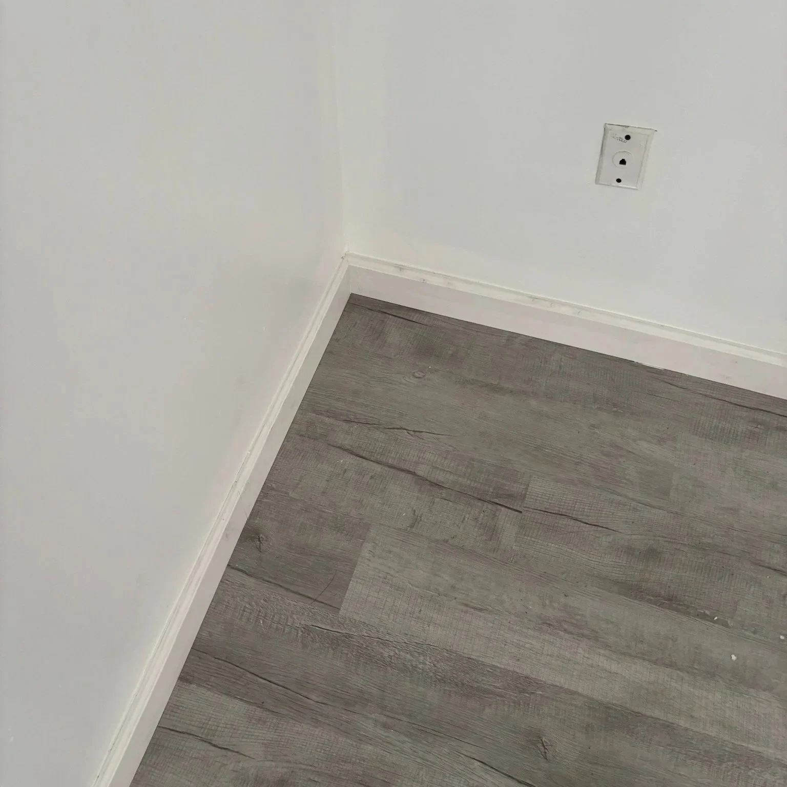 Image of a corner of a room with white walls, gray wooden floor, and an electrical outlet on the wall.