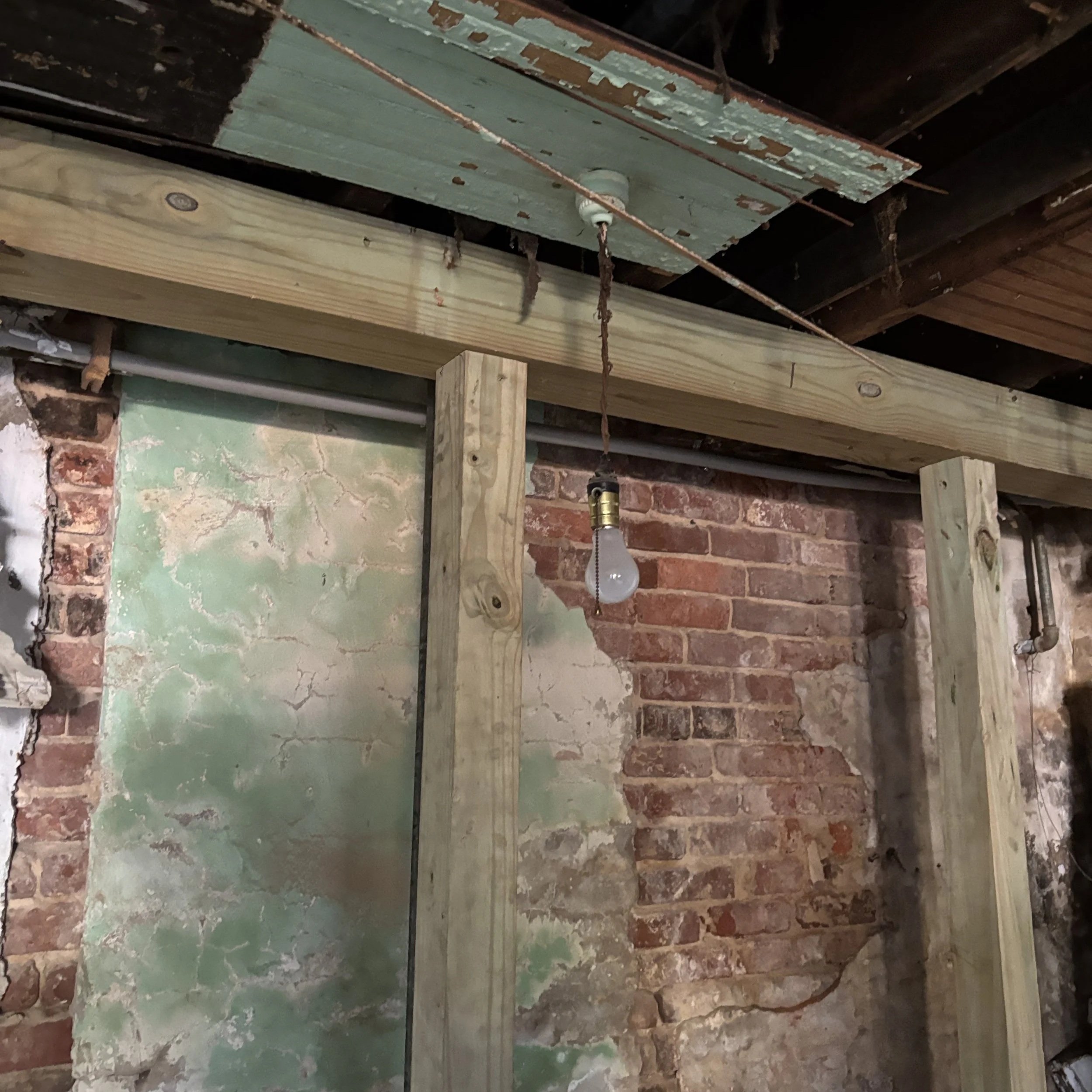 Exposed brick wall with peeling paint, wood framing, and a hanging light bulb in an unfinished industrial space.