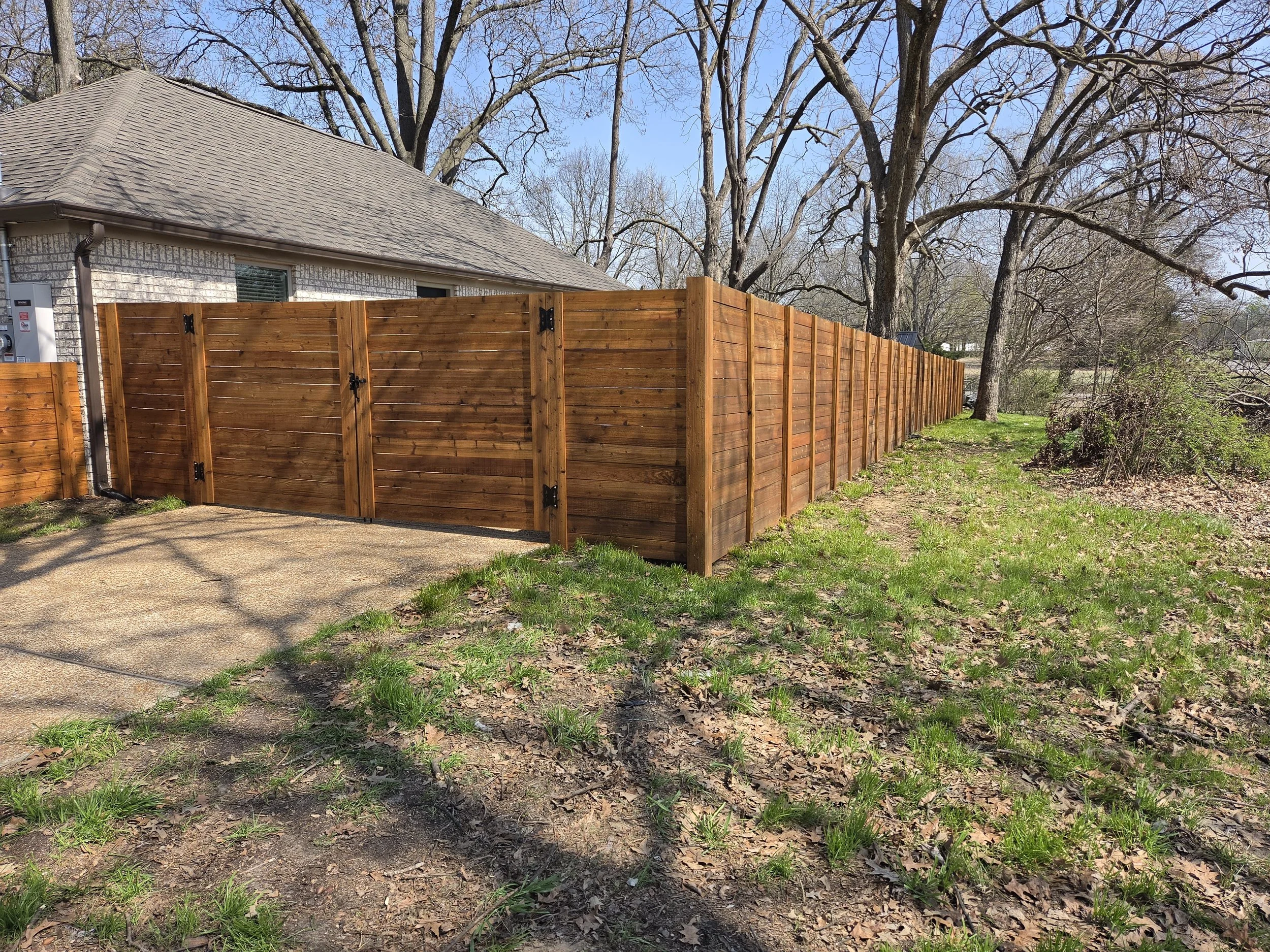 A wooden privacy fence surrounds the backyard of a house with a gray roof. The yard has patches of grass and bare soil with brown leaves, and of the trees, some are leafless, suggesting early spring.