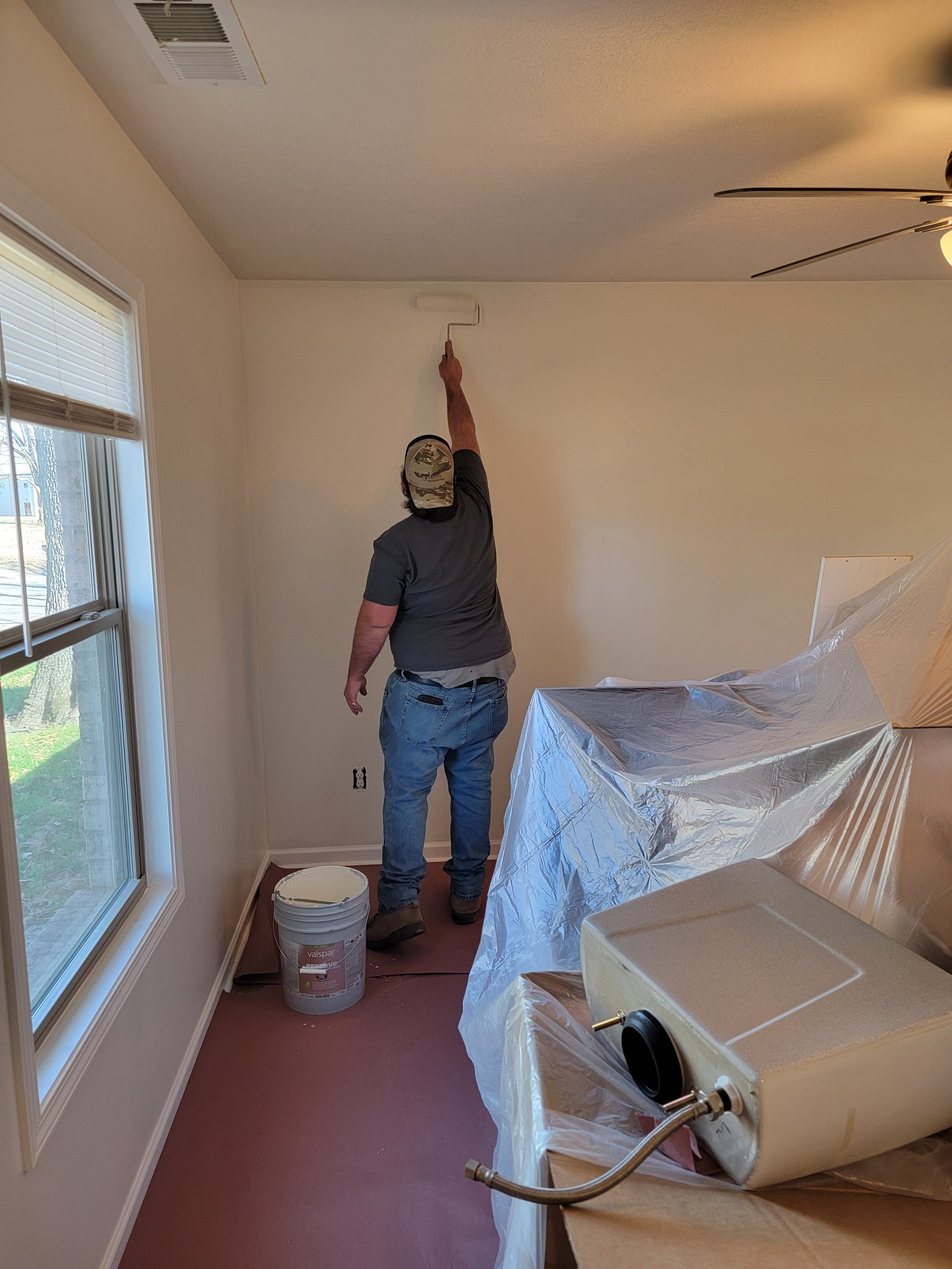 A person painting a ceiling with a paint roller in a room with window, plastic-covered furniture, and a bucket of paint on the floor.