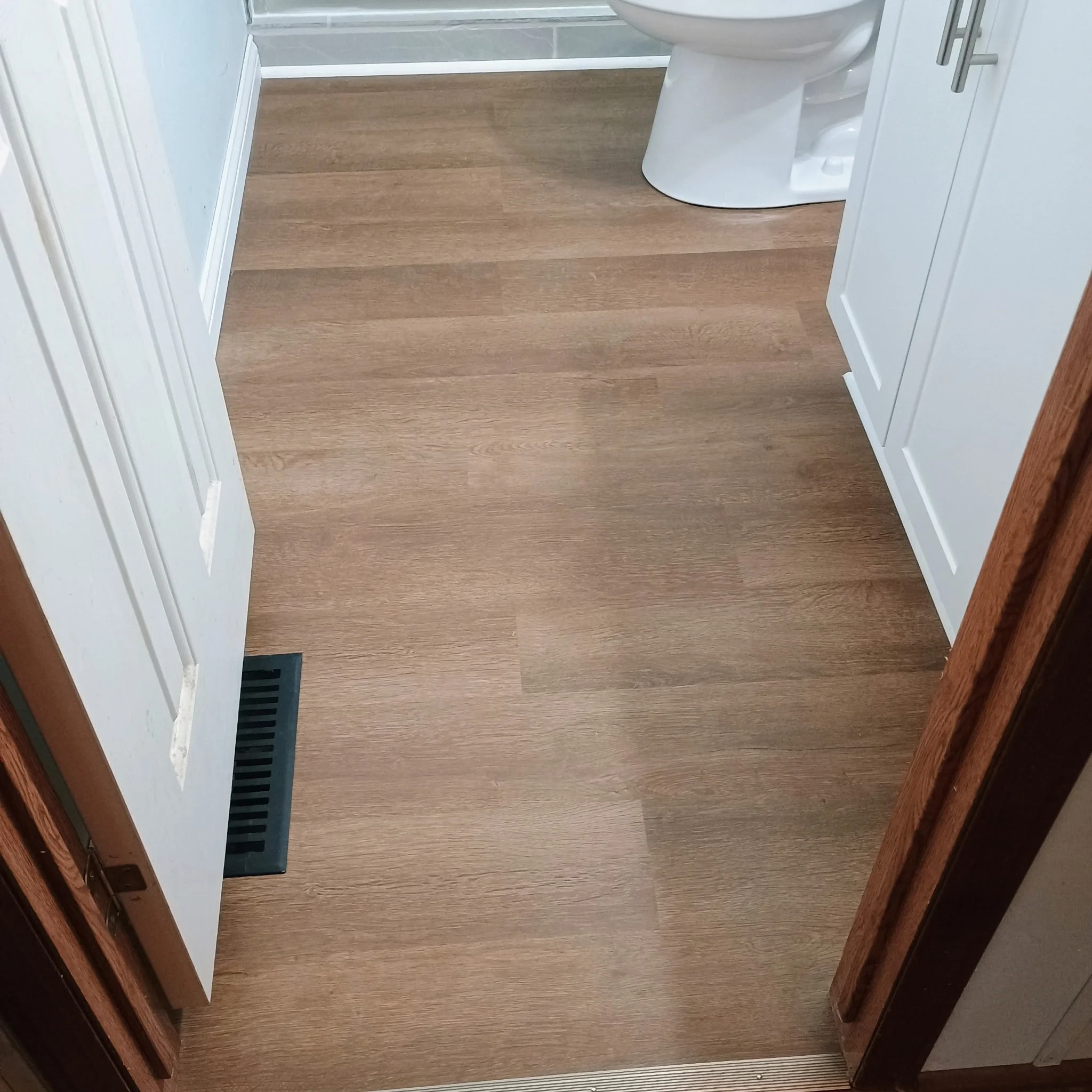 View of a bathroom floor with light brown wood-like flooring, a white toilet in the corner, a white cabinet on the right, and a metallic vent on the left side near the bottom.