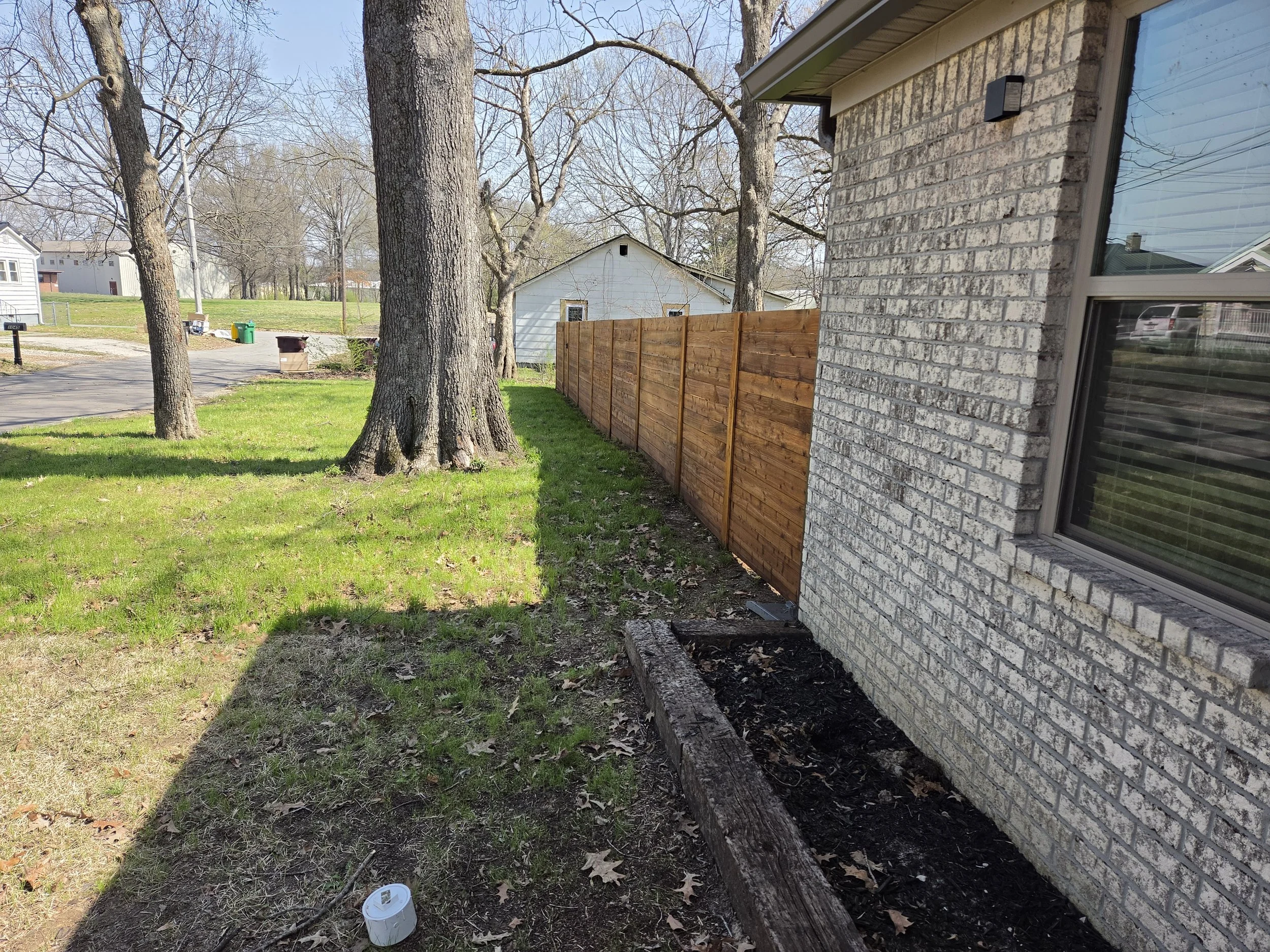 Side yard with fresh wooden fence, brick house wall, large tree, new flower bed, and patches of sunlight and shadow.