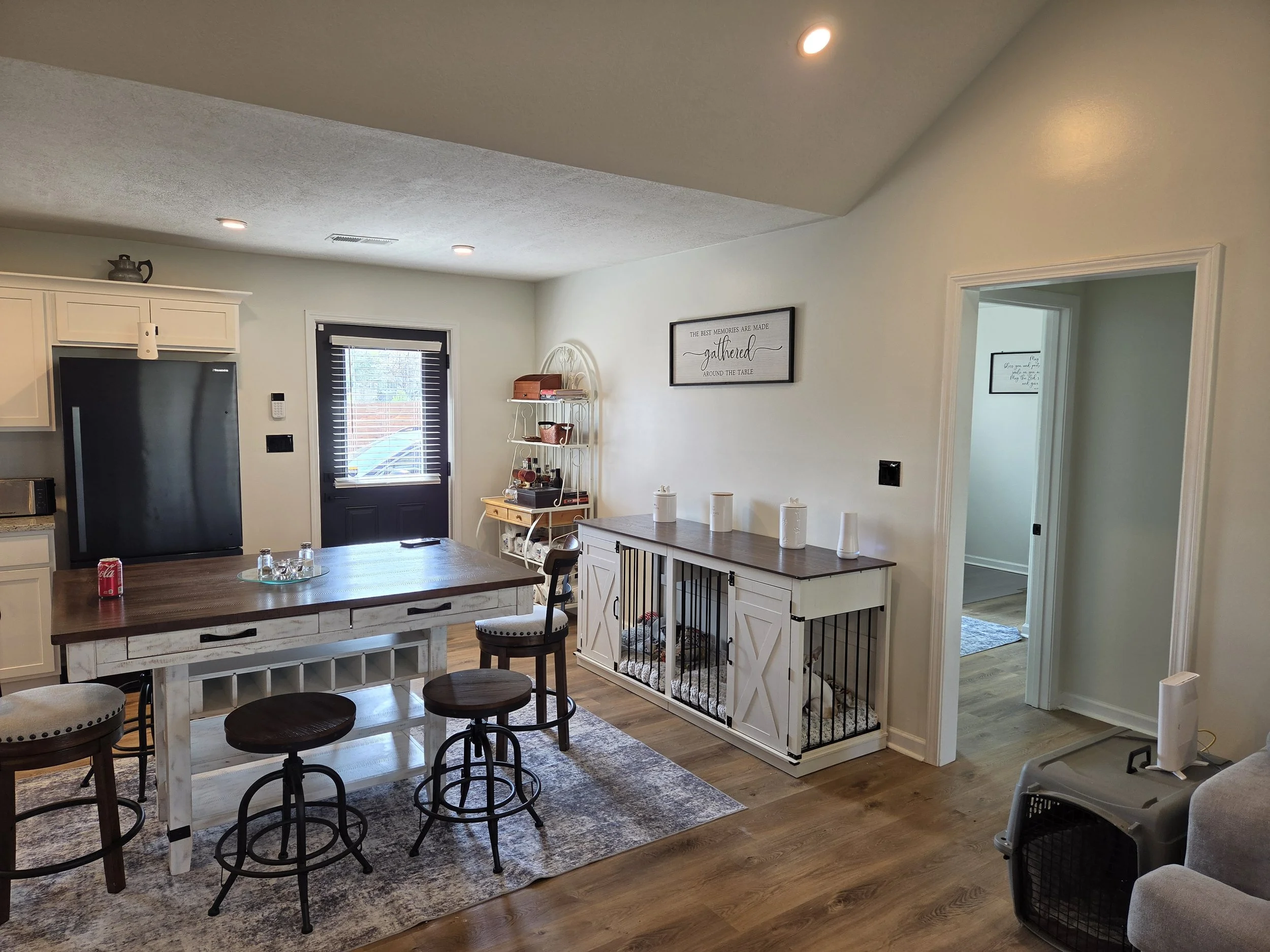 Living room with kitchen, wooden island, bar stools, white cabinetry, black door, wall art, shelving unit, dog crate, and carpeted pet area.