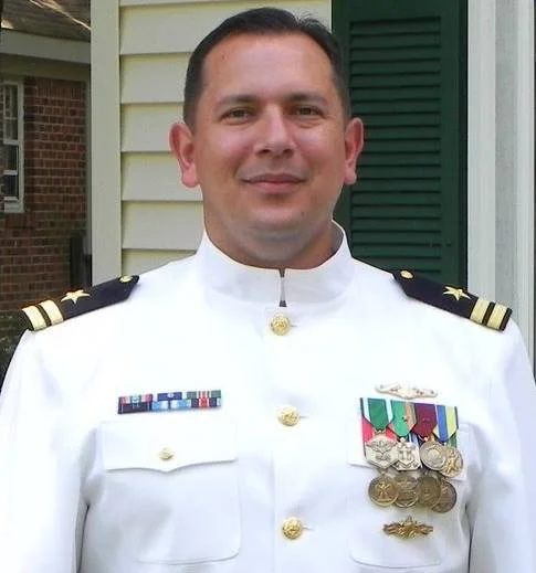 Jeremy James in a white military uniform with medals and insignia, standing outdoors in front of a yellow house.