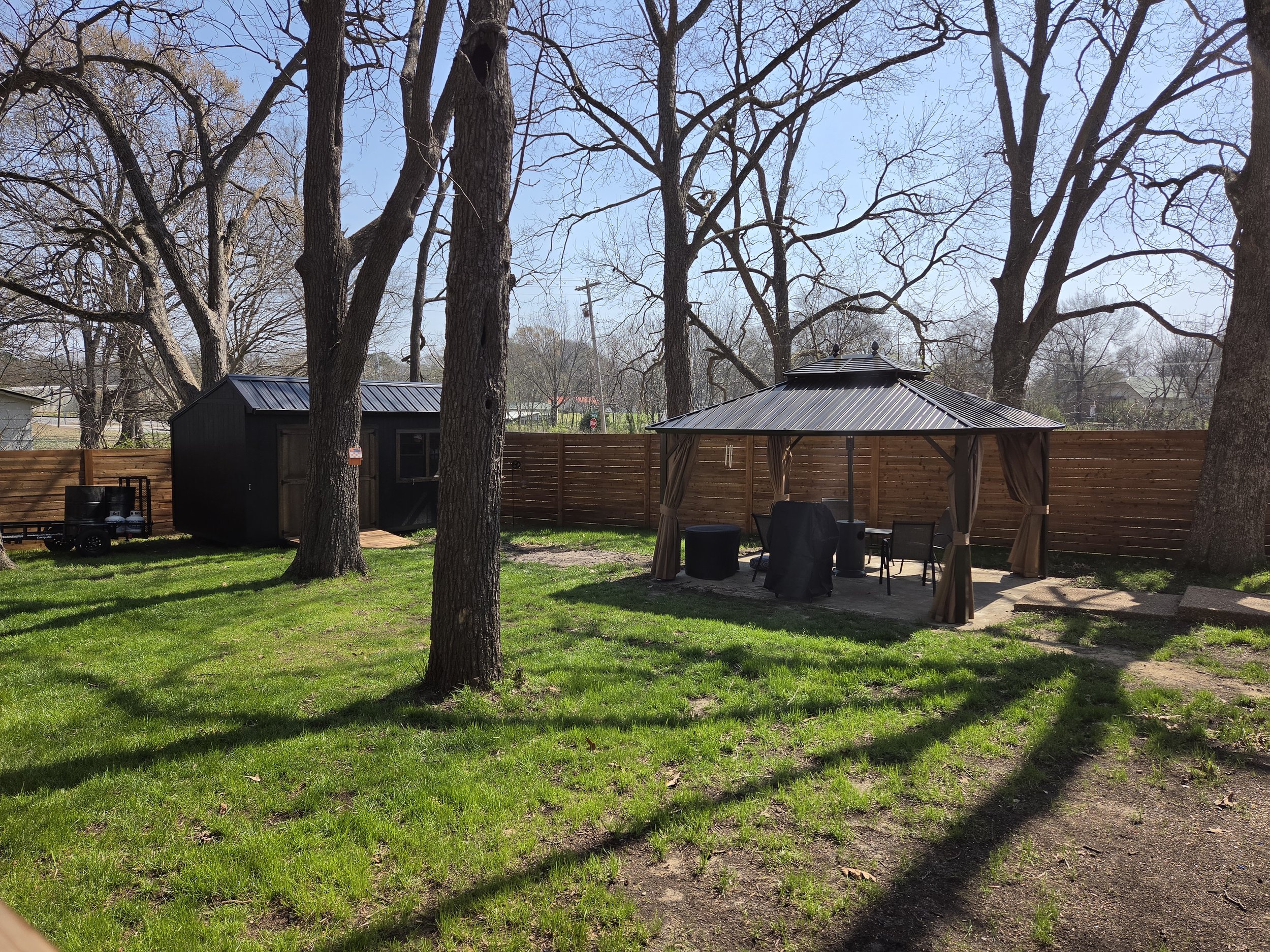 Backyard with green grass, several large leafless trees, a wooden fence, a black shed, and a patio gazebo with outdoor furniture.