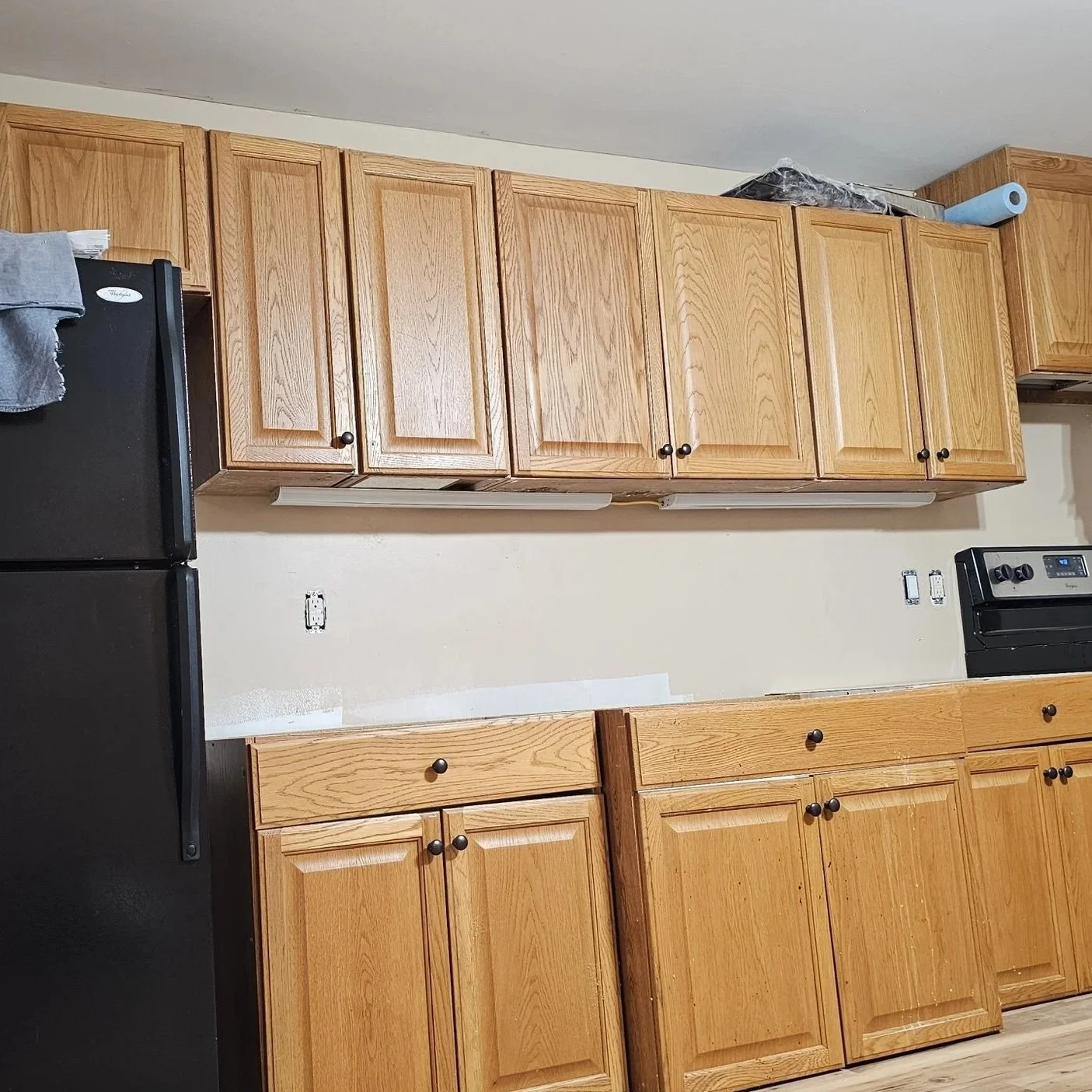 A kitchen with wooden cabinets, black refrigerator on the left, and black stove on the right, with an empty beige wall in the center.