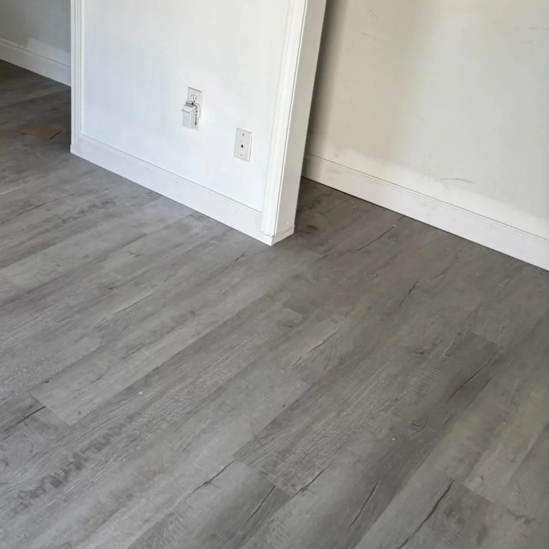 Interior corner with gray wood-look flooring, white walls, electrical outlets, and baseboards.