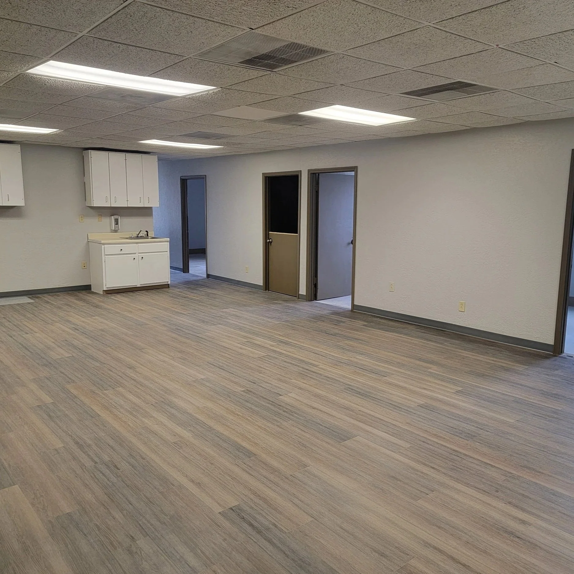 Empty room with wood-like flooring, white textured walls, white ceiling tiles, and fluorescent lights. There are three doorways on the back wall, one with a security screen door. On the left, there is a small white kitchenette area with white cabinets and a sink.