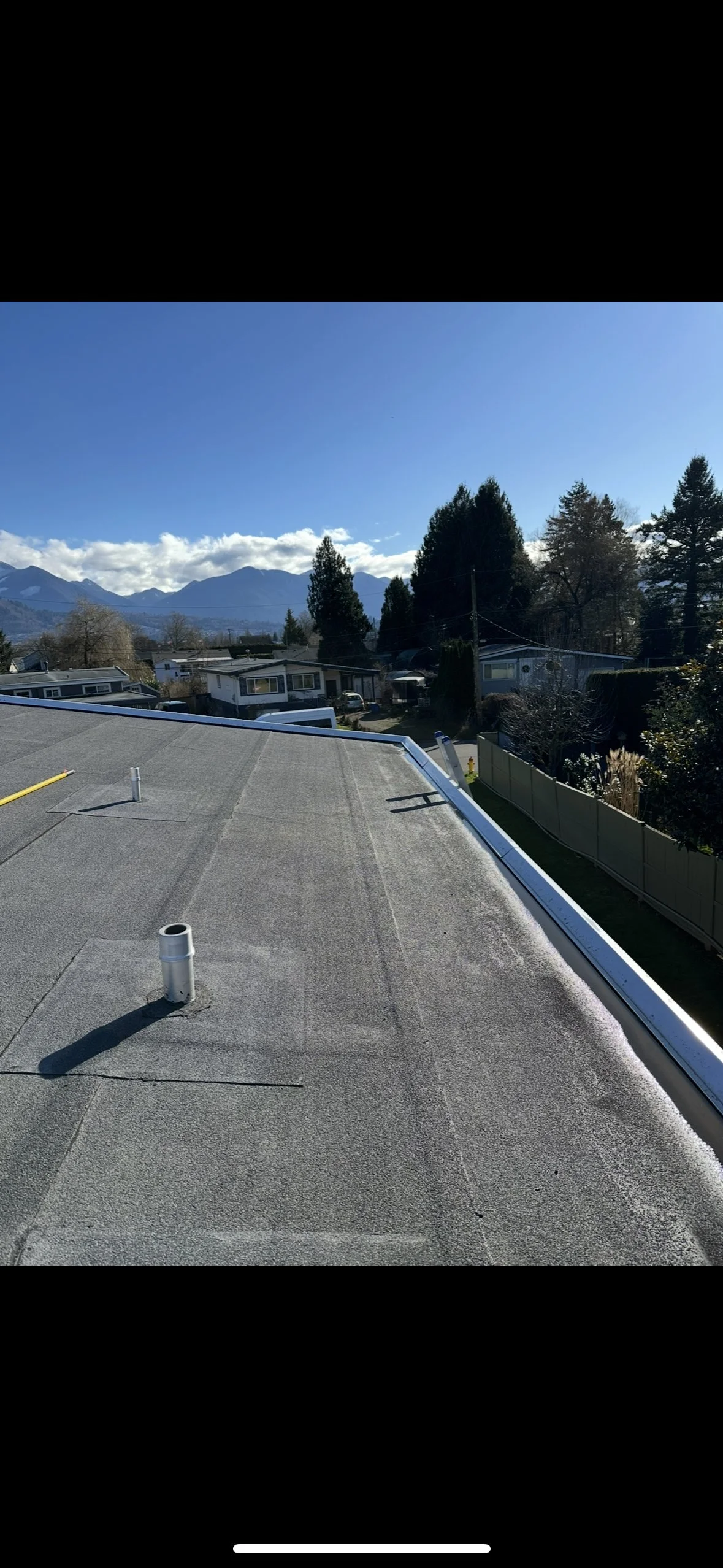 View from a rooftop showing a residential neighborhood with trees, houses, and mountains in the distance under a clear blue sky.