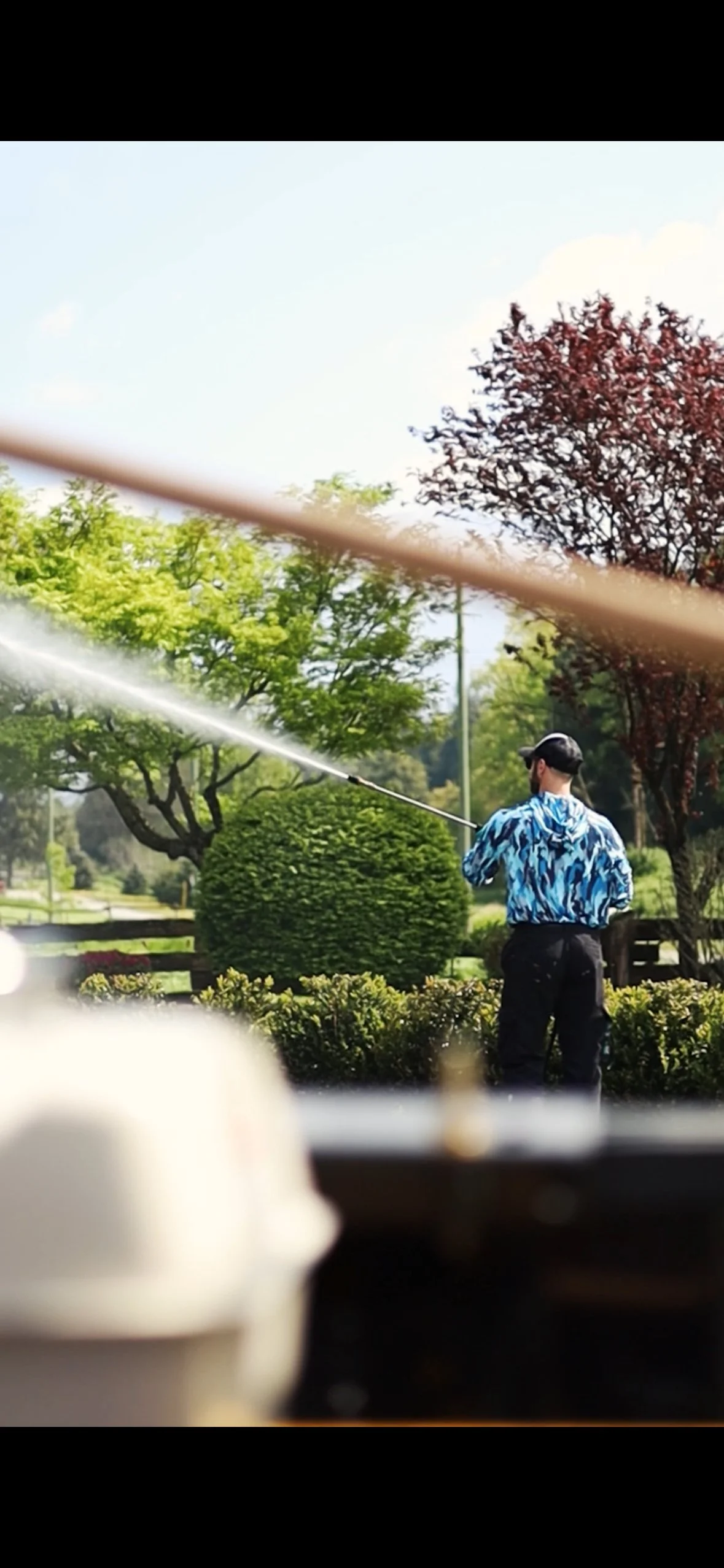 Person wearing a blue patterned jacket and black pants is watering plants in a park surrounded by trees with green and reddish leaves.