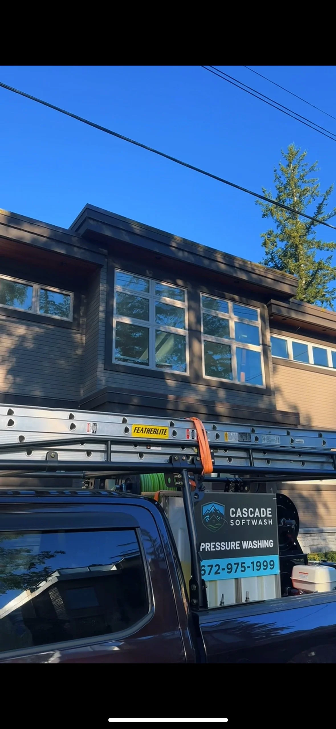 A house with large windows and wood siding, with a clear blue sky and a tall tree in the background. A truck with pressure washing equipment is parked in front.