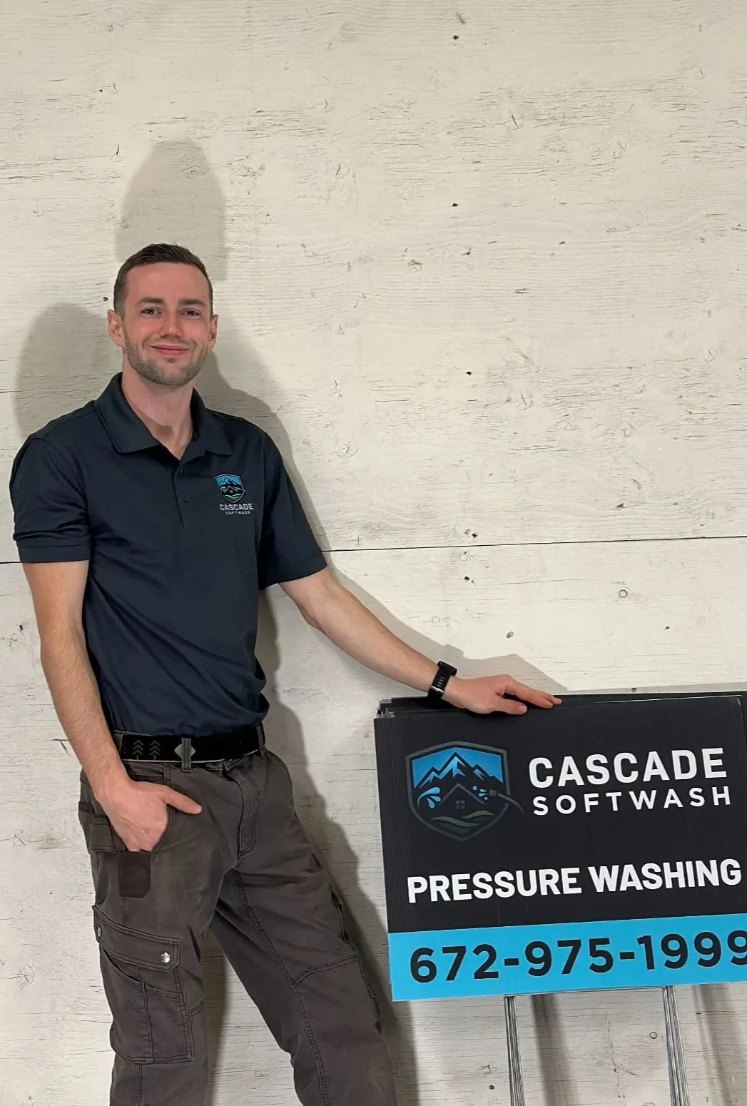 A man in a black Cascade Softwash polo shirt standing next to a sign that reads "Cascade Softwash Pressure Washing" with a mountain logo and a phone number.