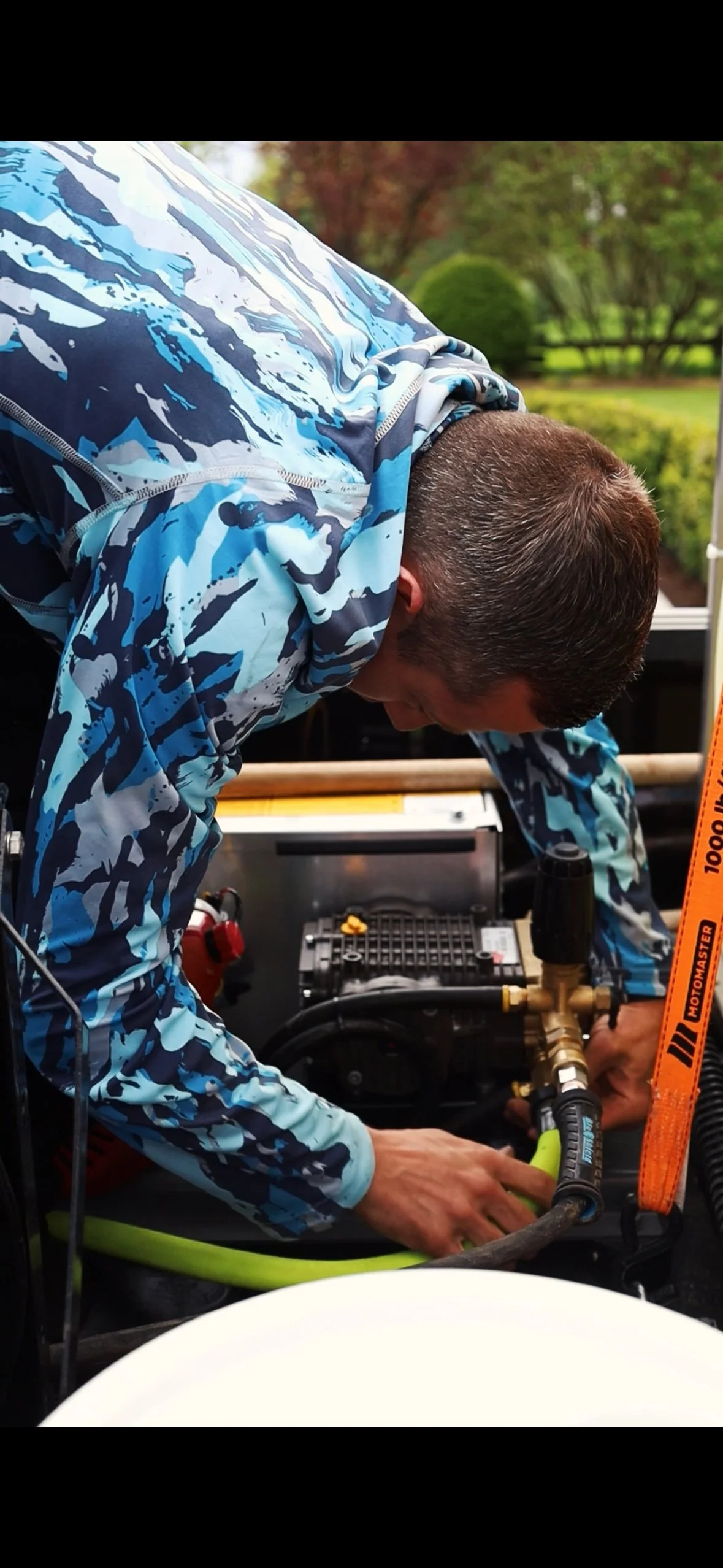 Man in blue camouflage jacket working on a piece of machinery outdoors, with trees and green foliage in the background.