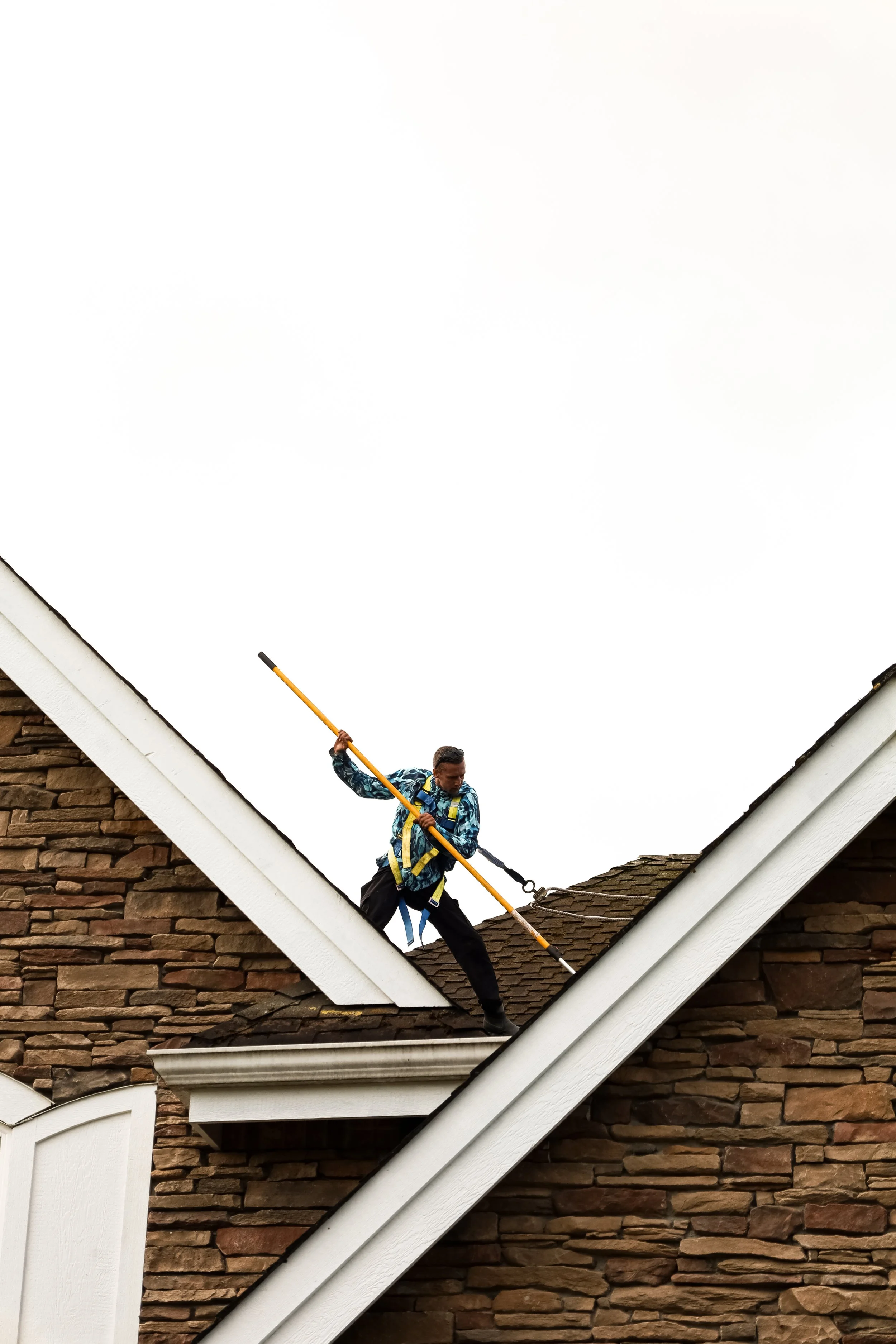 A man wearing safety harness and work gear cleaning a roof with a long pole on a house with brick exterior and white trim.
