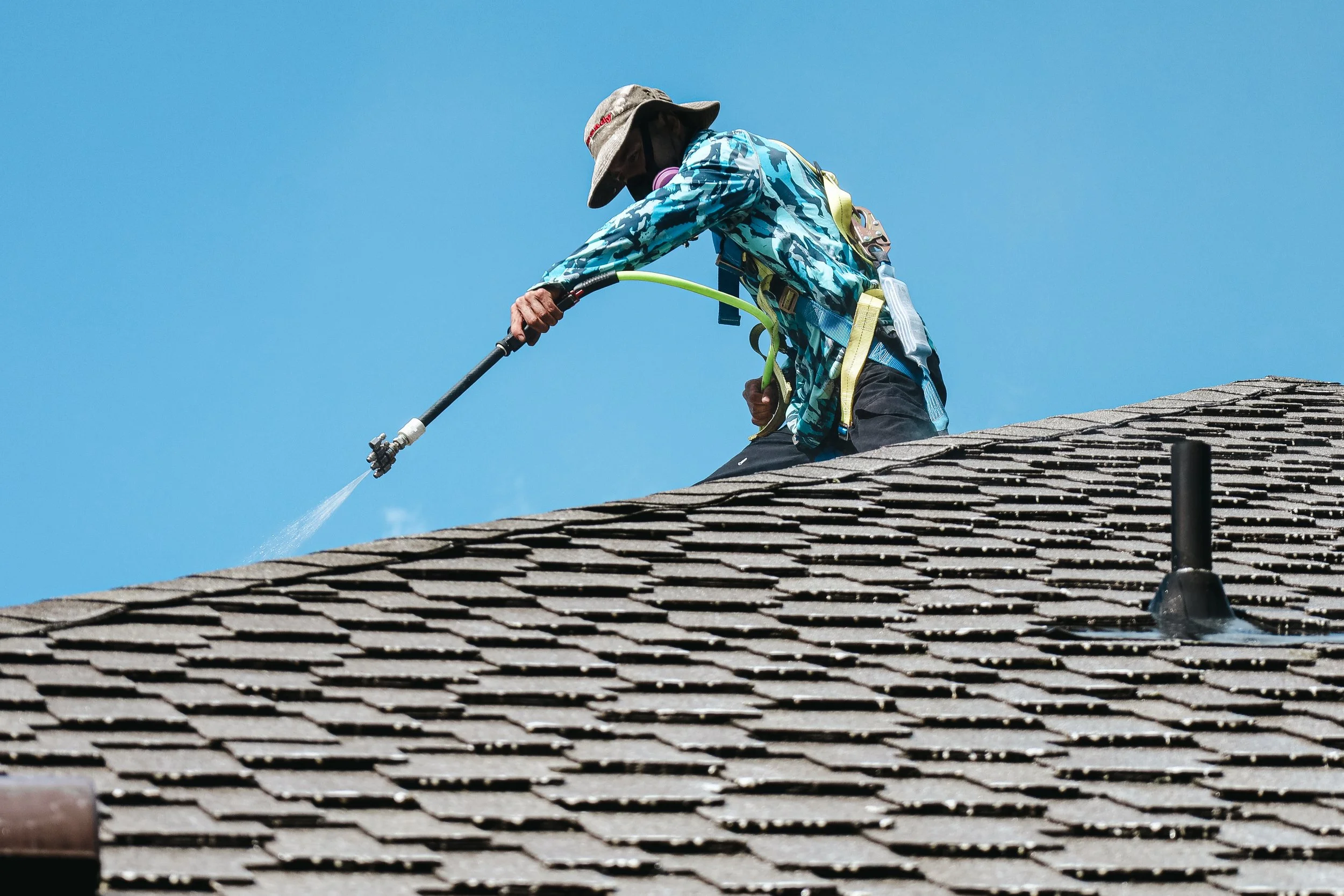 A person wearing a wide-brimmed hat and a camouflage jacket is on a rooftop, using a hose to spray water on the shingles under a clear blue sky.
