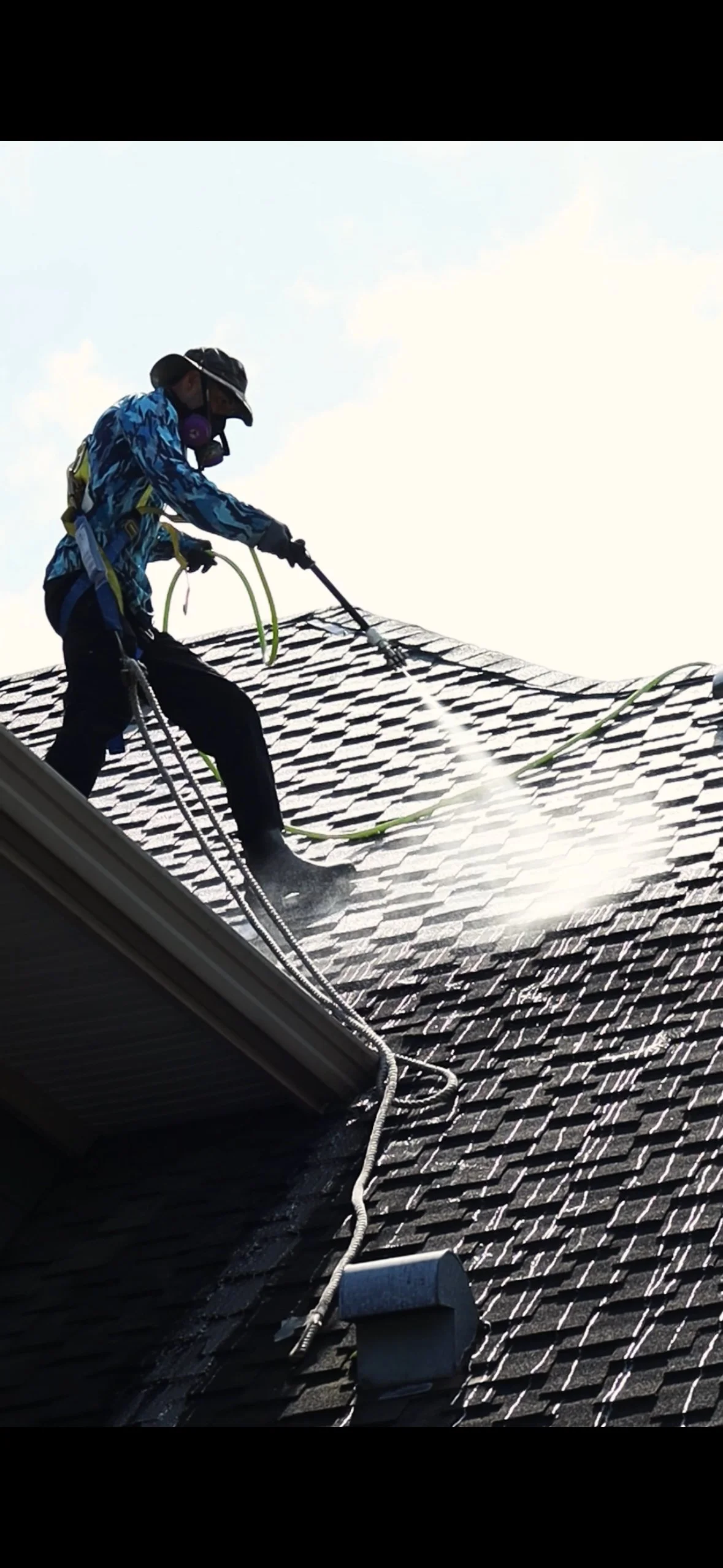 A person wearing safety gear, including a helmet and mask, is cleaning a rooftop with a power washer.