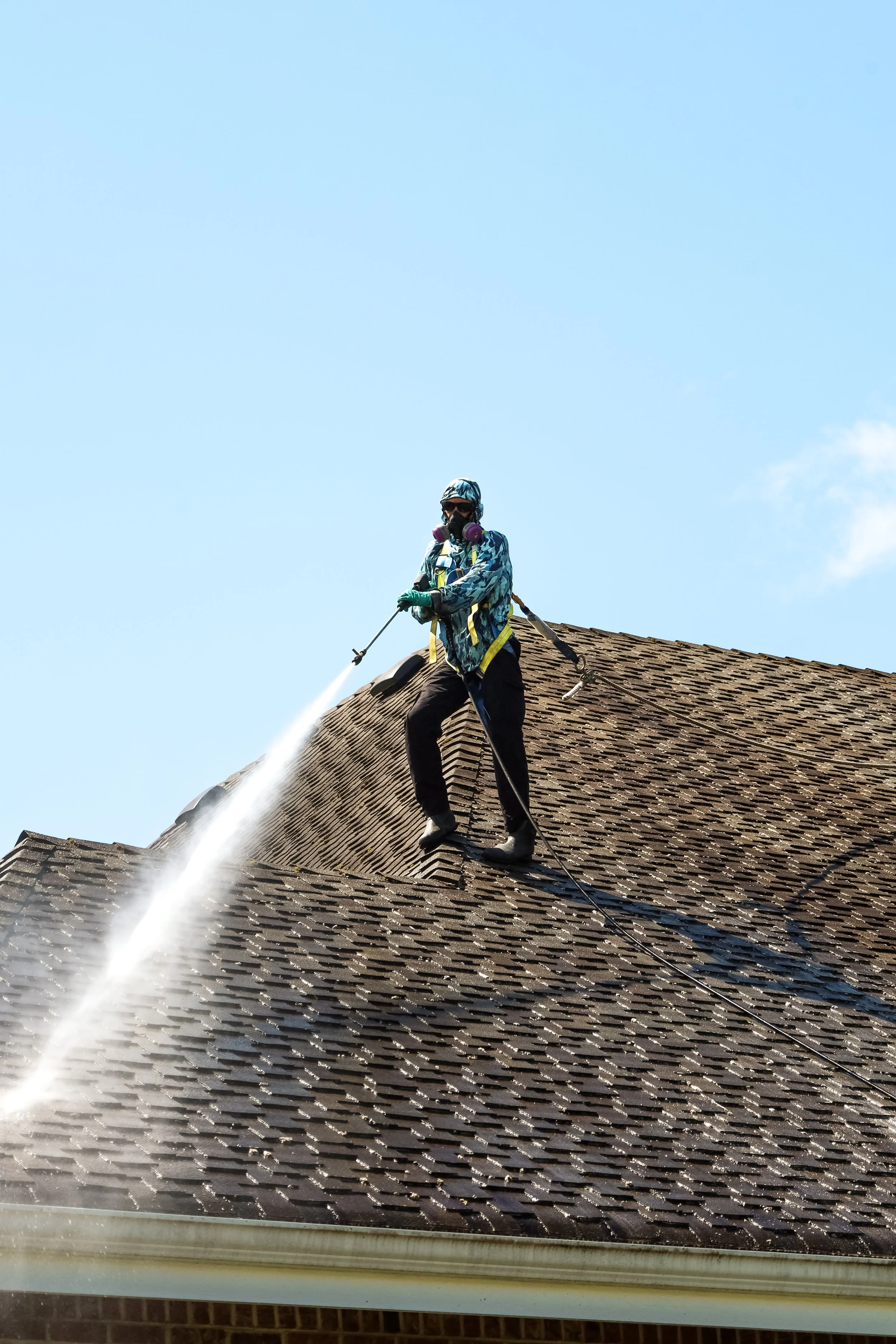 Person power washing a roof with a spray hose, wearing a hat, sunglasses, mask, and gloves, standing on the roof shingles.