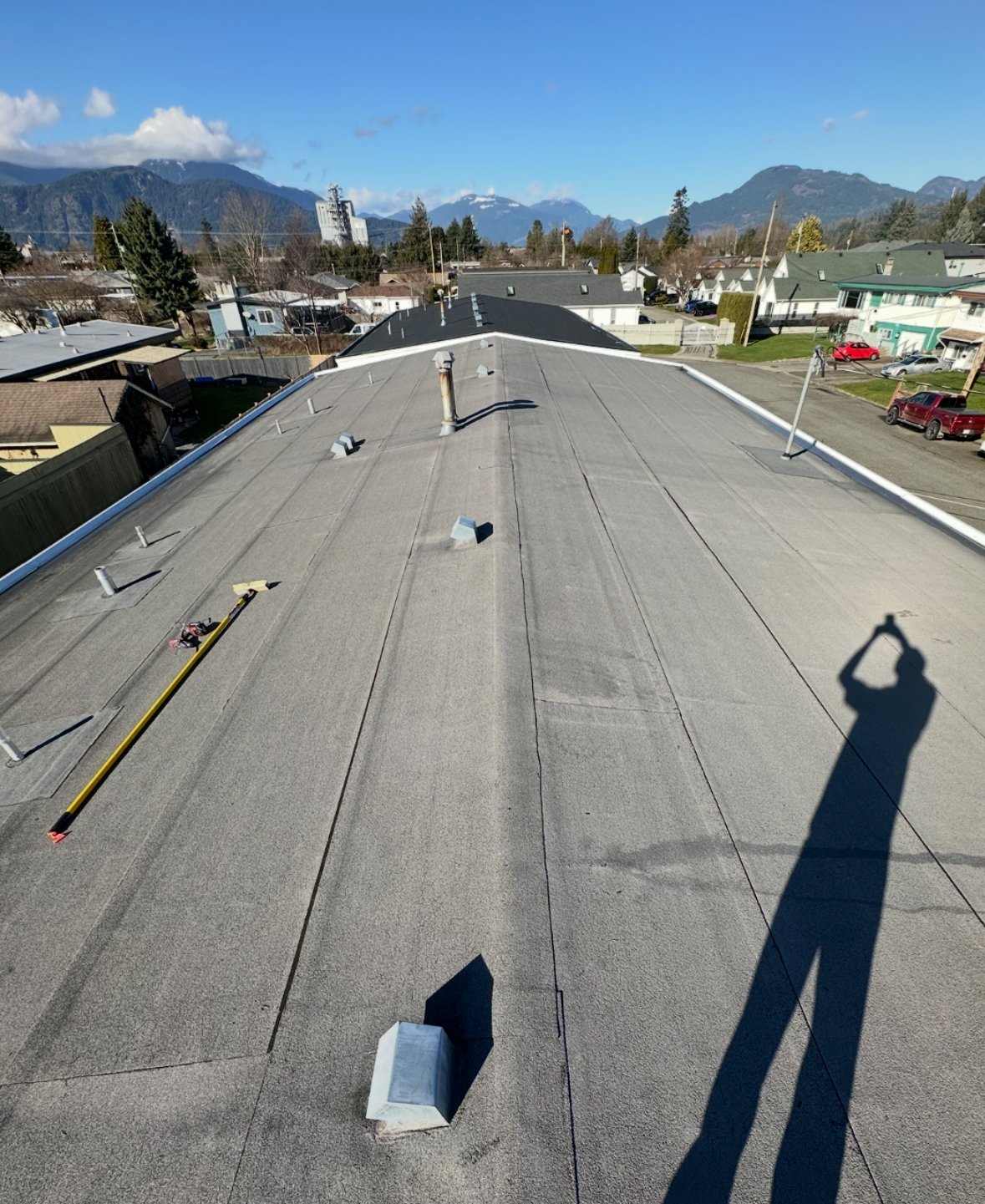 View of a rooftop in a residential neighborhood with mountains in the background. The roof has various vents and a shadow of a person holding a tool.