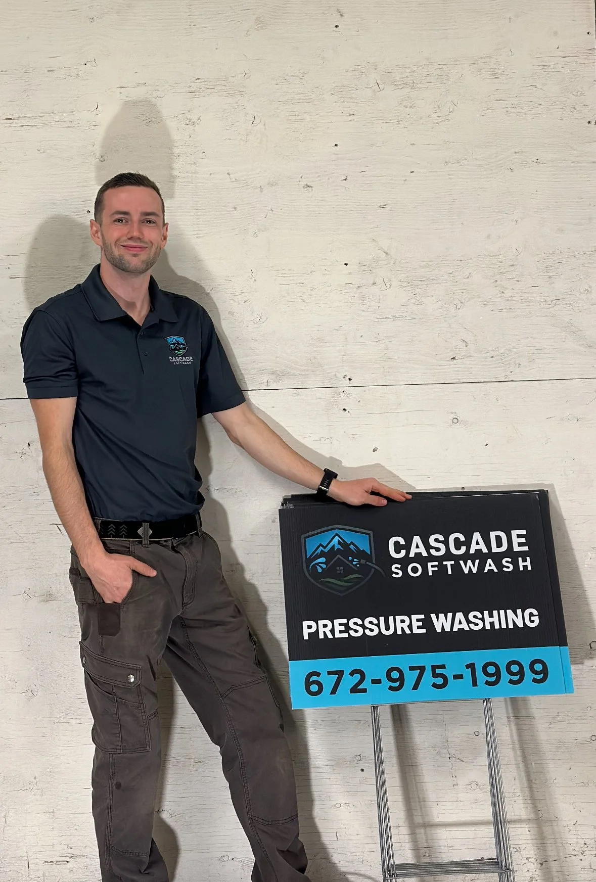 Man in dark polo shirt and cargo pants standing next to a sign for Cascade Softwash, promoting pressure washing services, with a white wooden background.