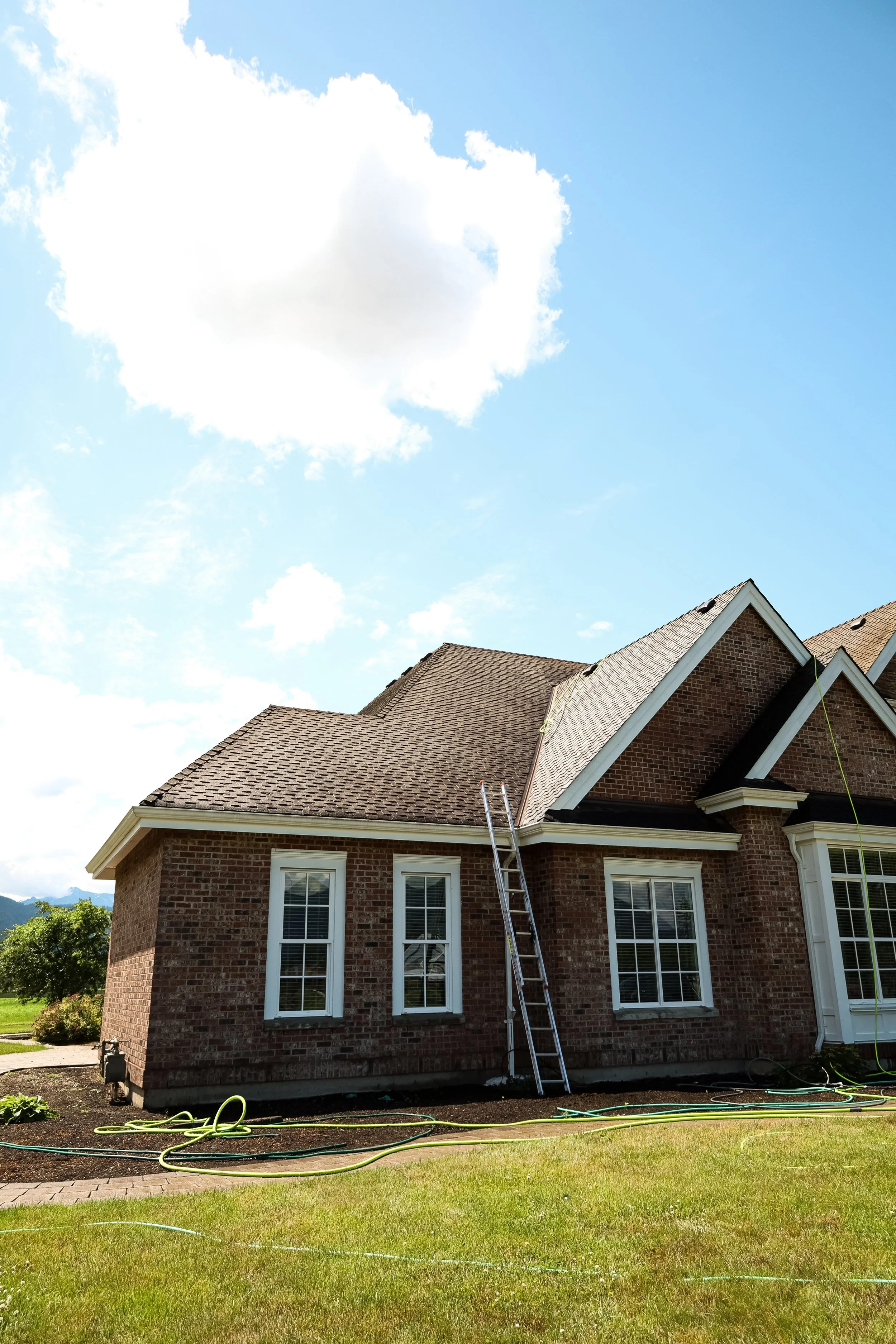 A brick house with a gabled roof and tall windows, a ladder leaning against the side, with a blue sky and white clouds overhead.