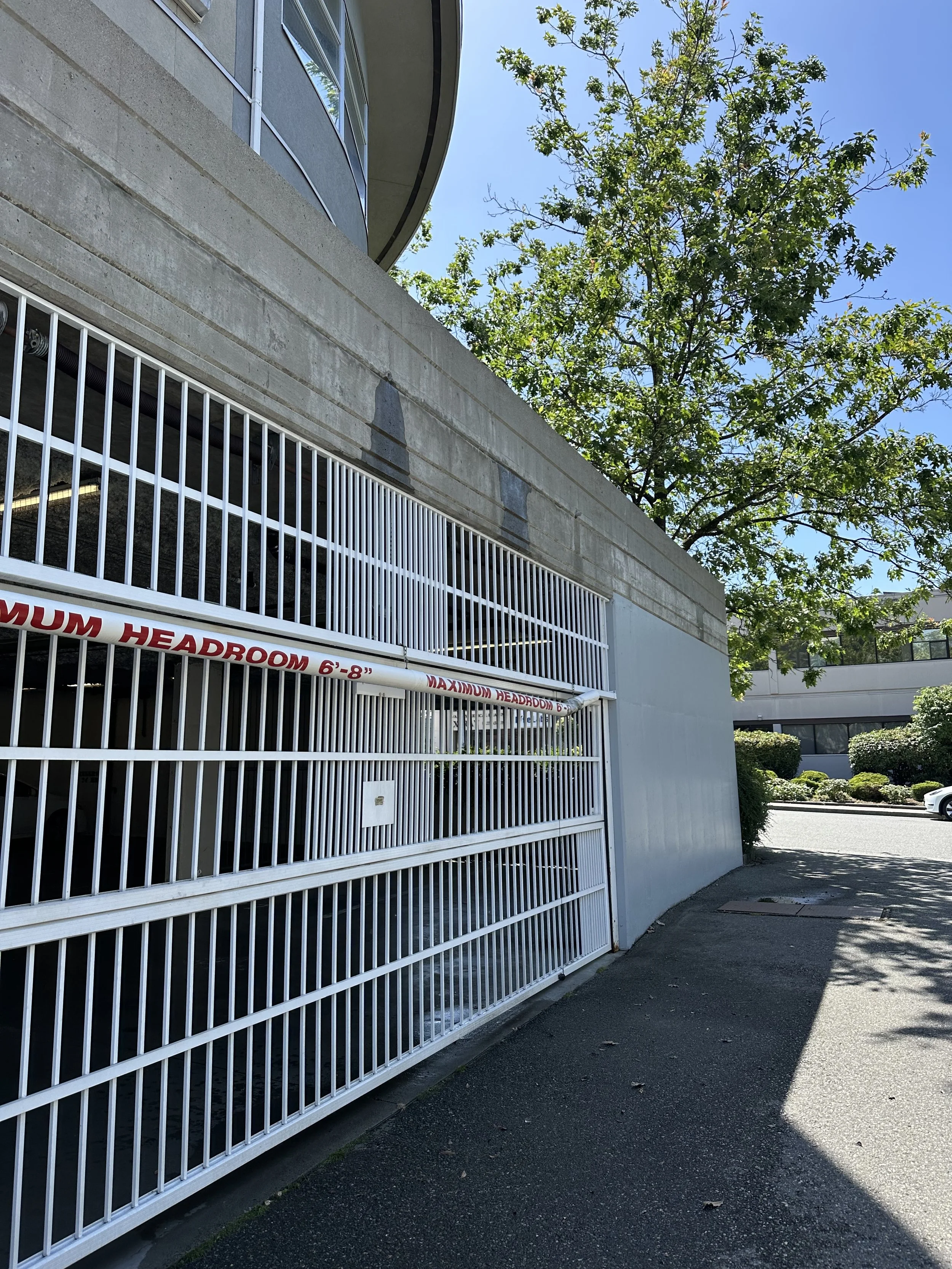 A white metal gate with a sign that reads 'MAXIMUM HEADROOM 6'-8"' in red letters, attached to a concrete building, with a tree and a clear blue sky in the background.