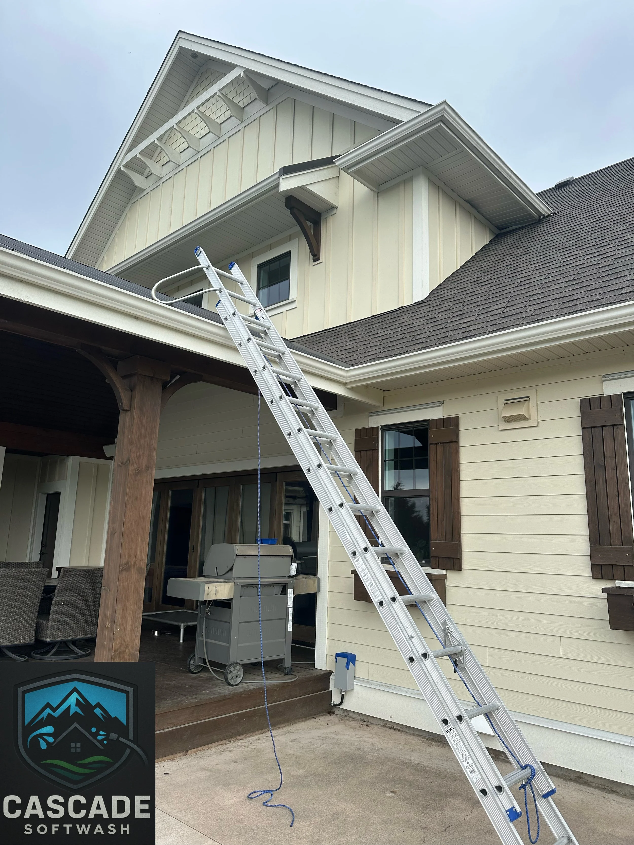 Ladder leaning against the second story of a house with beige siding and dark brown shutters, with a covered porch area below and patio furniture nearby. The house has a well-maintained roof with brown shingles, and there is a sign in the lower left 