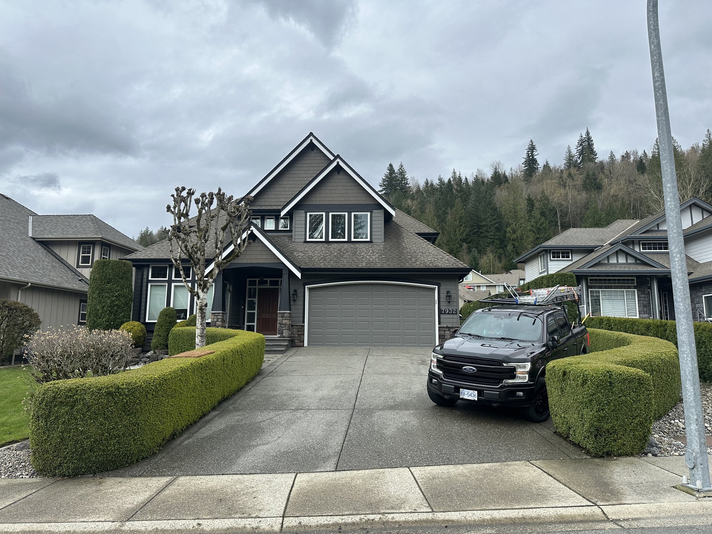 Modern gray two-story house with a front yard, neatly trimmed hedges, and an SUV parked on the driveway. The house has a garage, a small tree in the front yard, and trees on a hill in the background under cloudy skies.