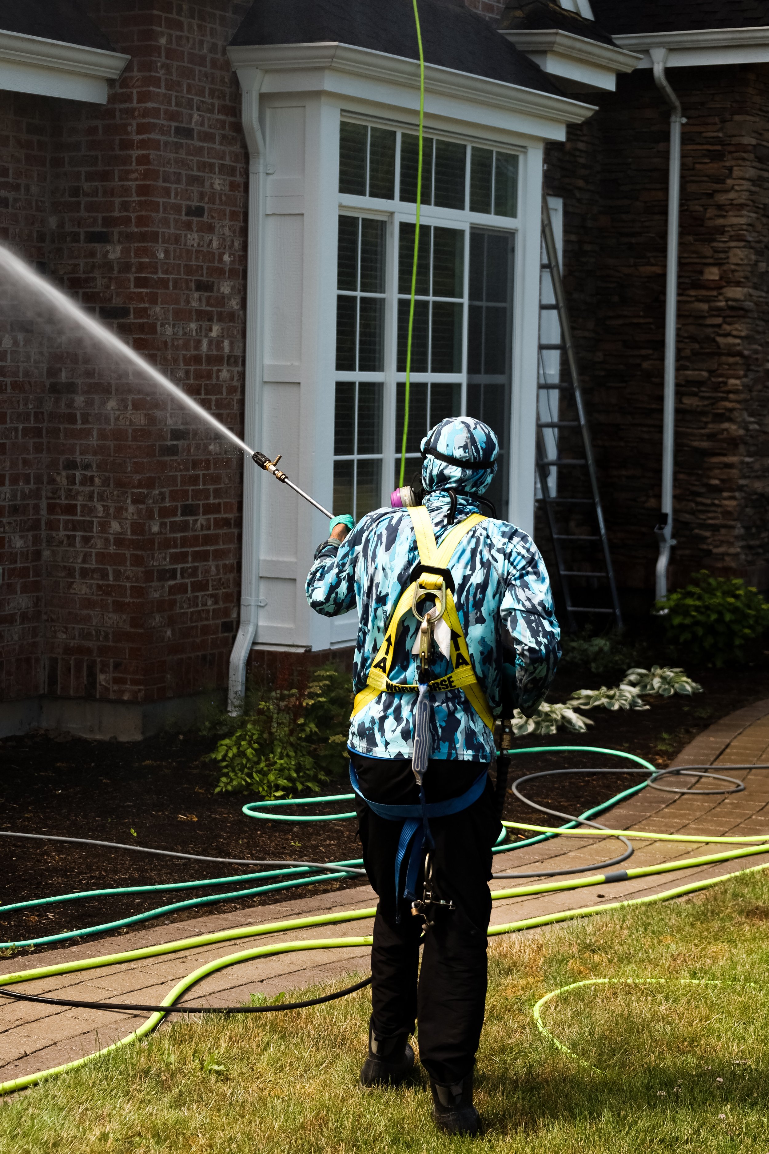 Person in camouflage clothing and safety gear spraying water on a house exterior, with hoses on the ground, near a brick and stone house with a large window.