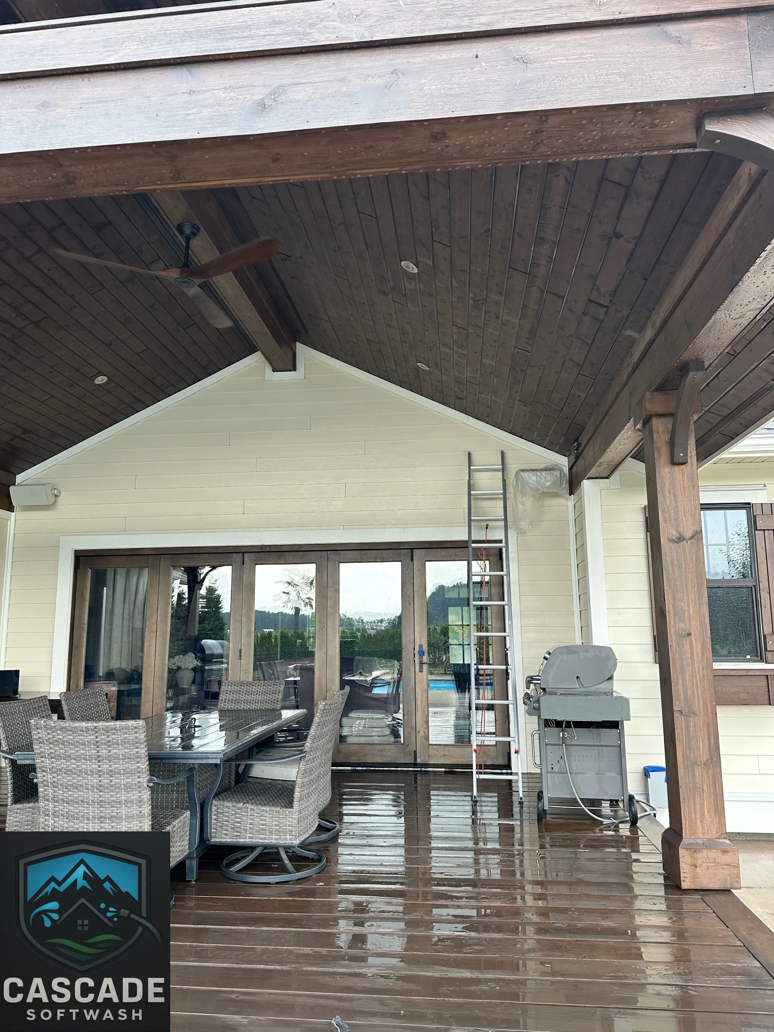 A covered outdoor patio area with wet wooden flooring, a dining table with wicker chairs, a gas grill, a ladder, and a ceiling fan, with a house and sliding glass door leading to the interior.