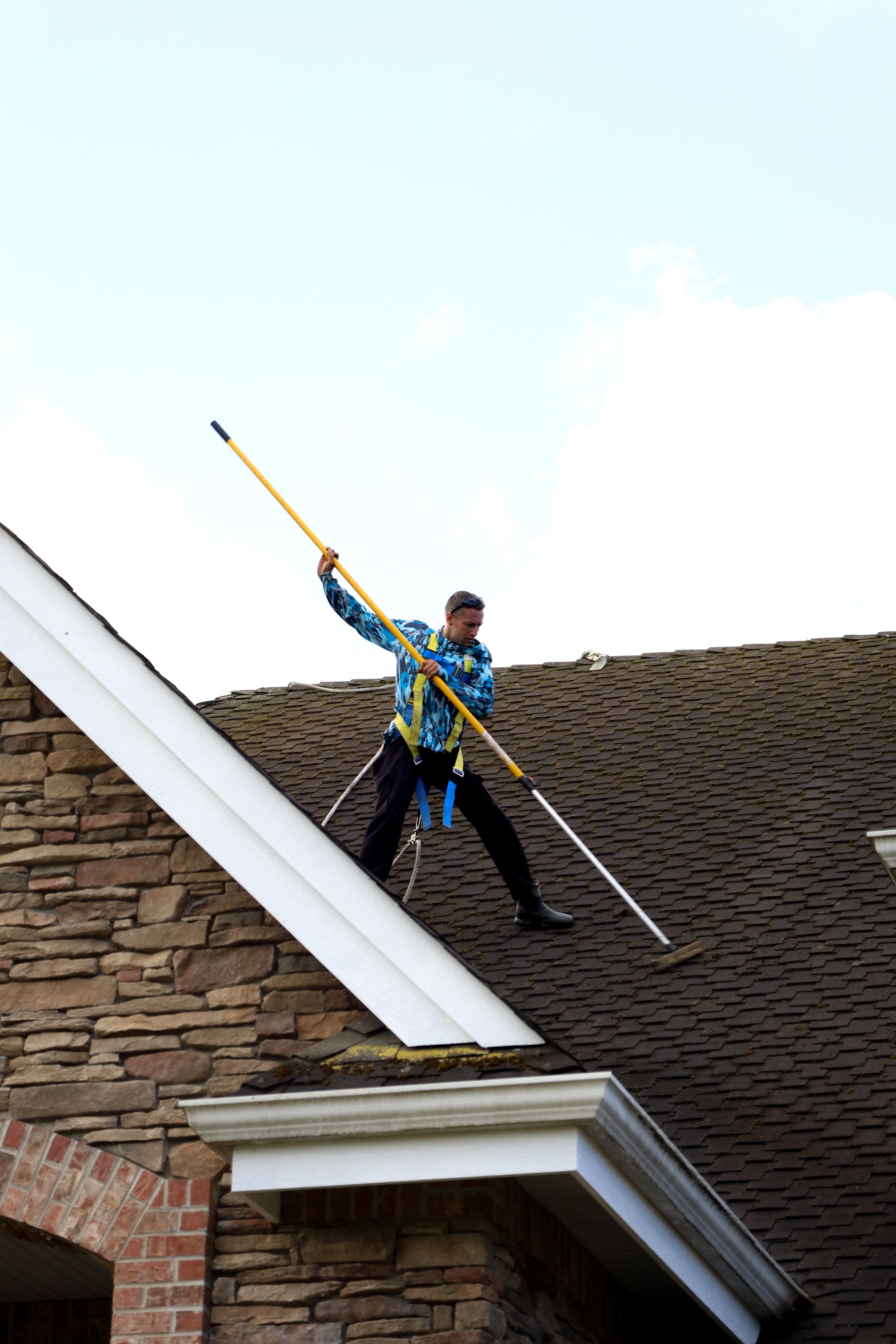 A person is standing on the roof of a house, holding a long pole and appears to be cleaning or inspecting the roof. The individual is wearing a blue patterned jacket, black pants, and rubber boots. The house has a brick and stone façade with a shingl