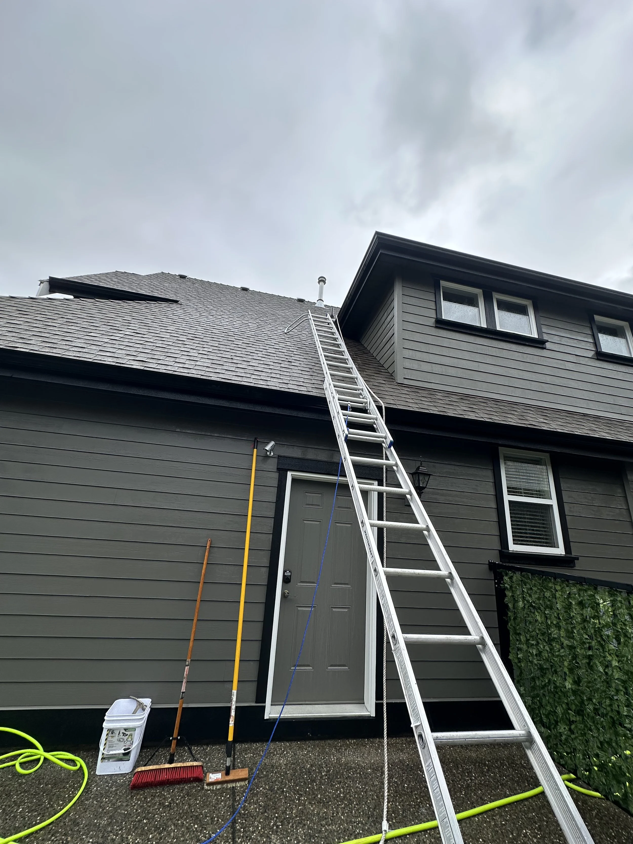 Ladder leaning against a gray house with a dark gray door, with cleaning tools and hoses on the ground, and a cloudy sky above.