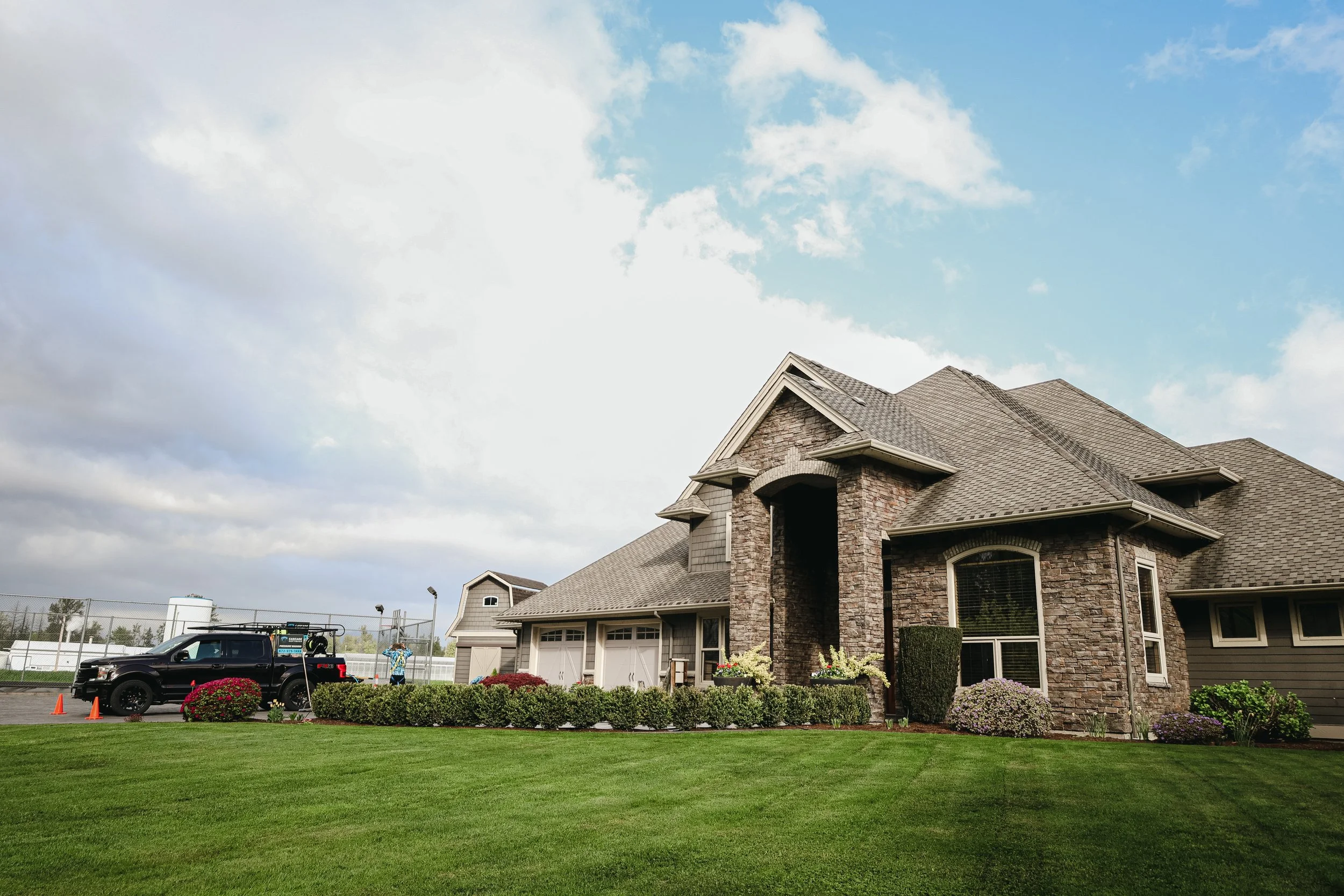 A large brick house with a steep roof, dormer windows, and a two-car garage. The front yard has green grass, potted plants, and bushes. There are cars and cones in the driveway, with a person standing near a parking meter. The sky is partly cloudy.