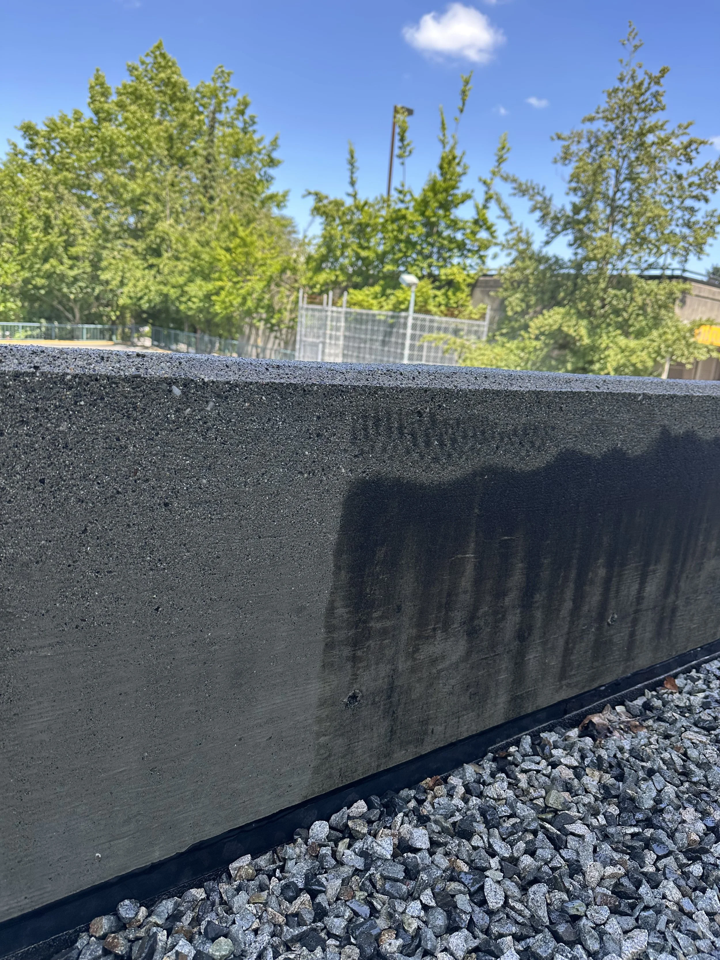 Close-up of a concrete barrier with a rough texture, with multicolored gravel underneath, and a background of lush green trees and a clear blue sky.