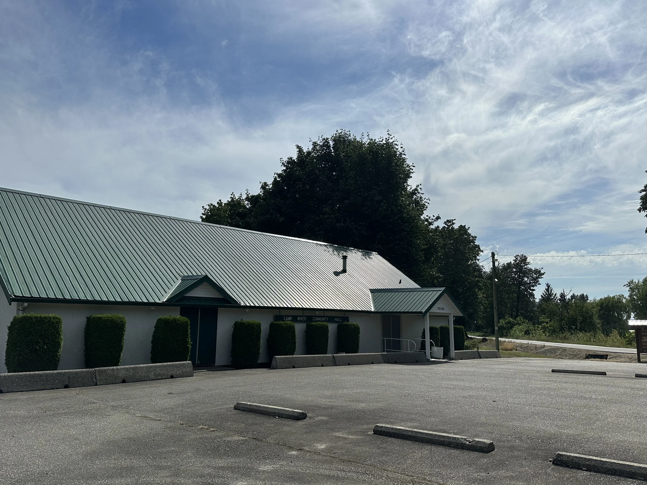 A building with a green metal roof and a sign reading 'Camp River Community Hall' in front of a small parking lot with concrete wheel stops. There are neatly trimmed bushes along the building and a large tree behind it. The sky is partly cloudy with 