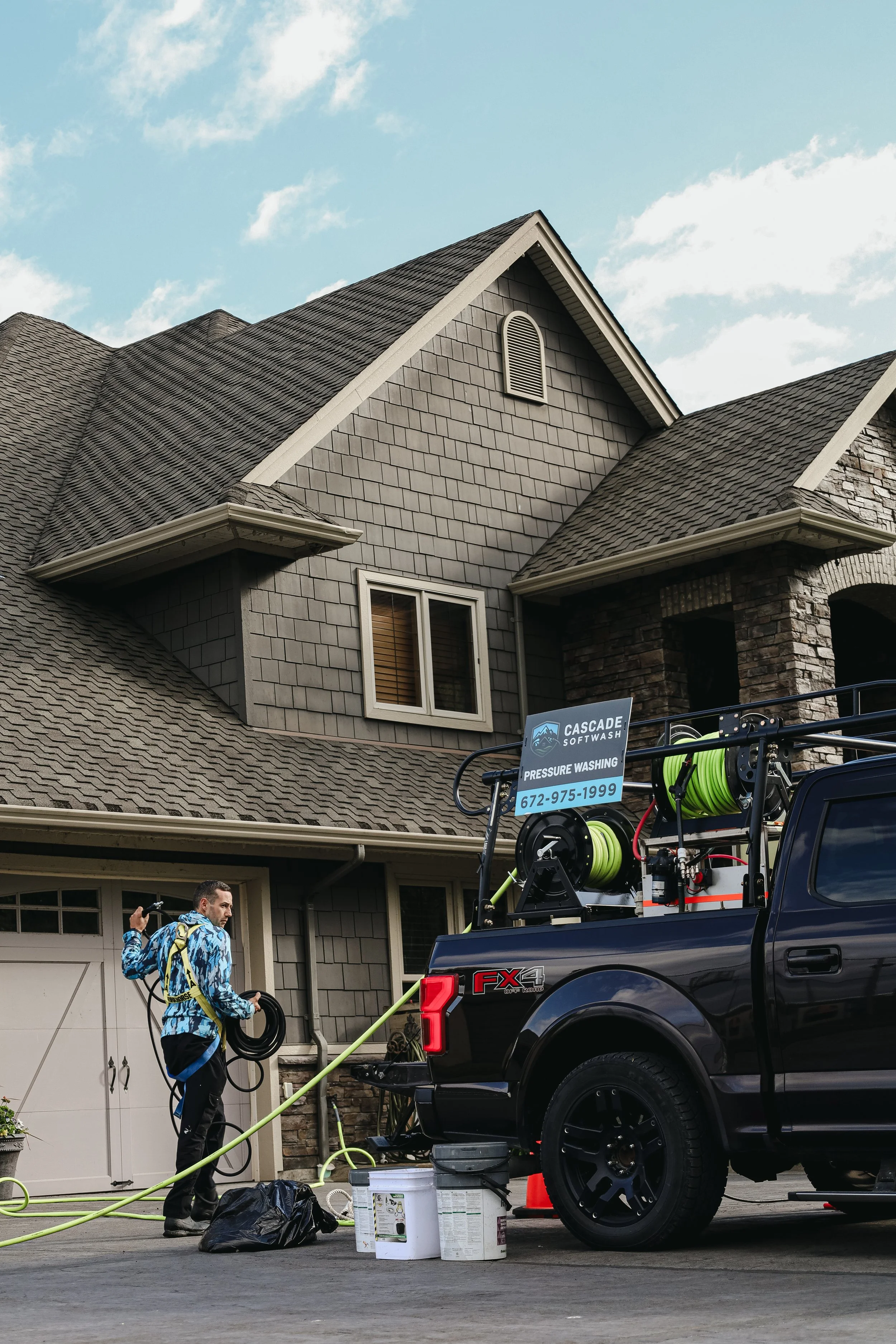 A man in a blue patterned outfit carrying a hose and equipment stands in front of a black pickup truck with someone washing a house. There's a sign on the truck for a pressure washing service. The house has a gray shingled roof, a small window, and a