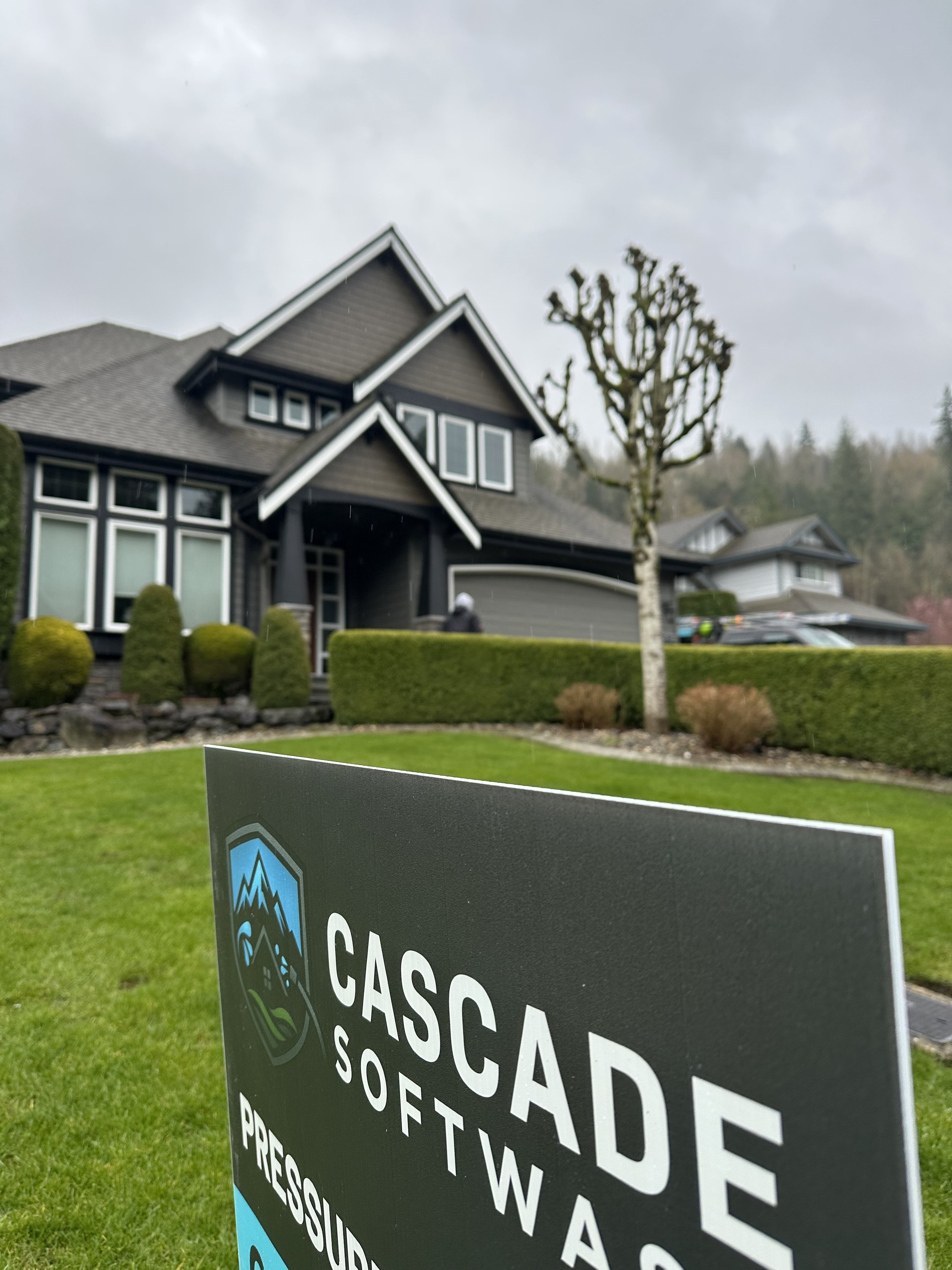 A house with modern architecture and dark siding, surrounded by well-maintained lawn and bushes, with a sign in the foreground reading 'Cascades Software Headquarters'.
