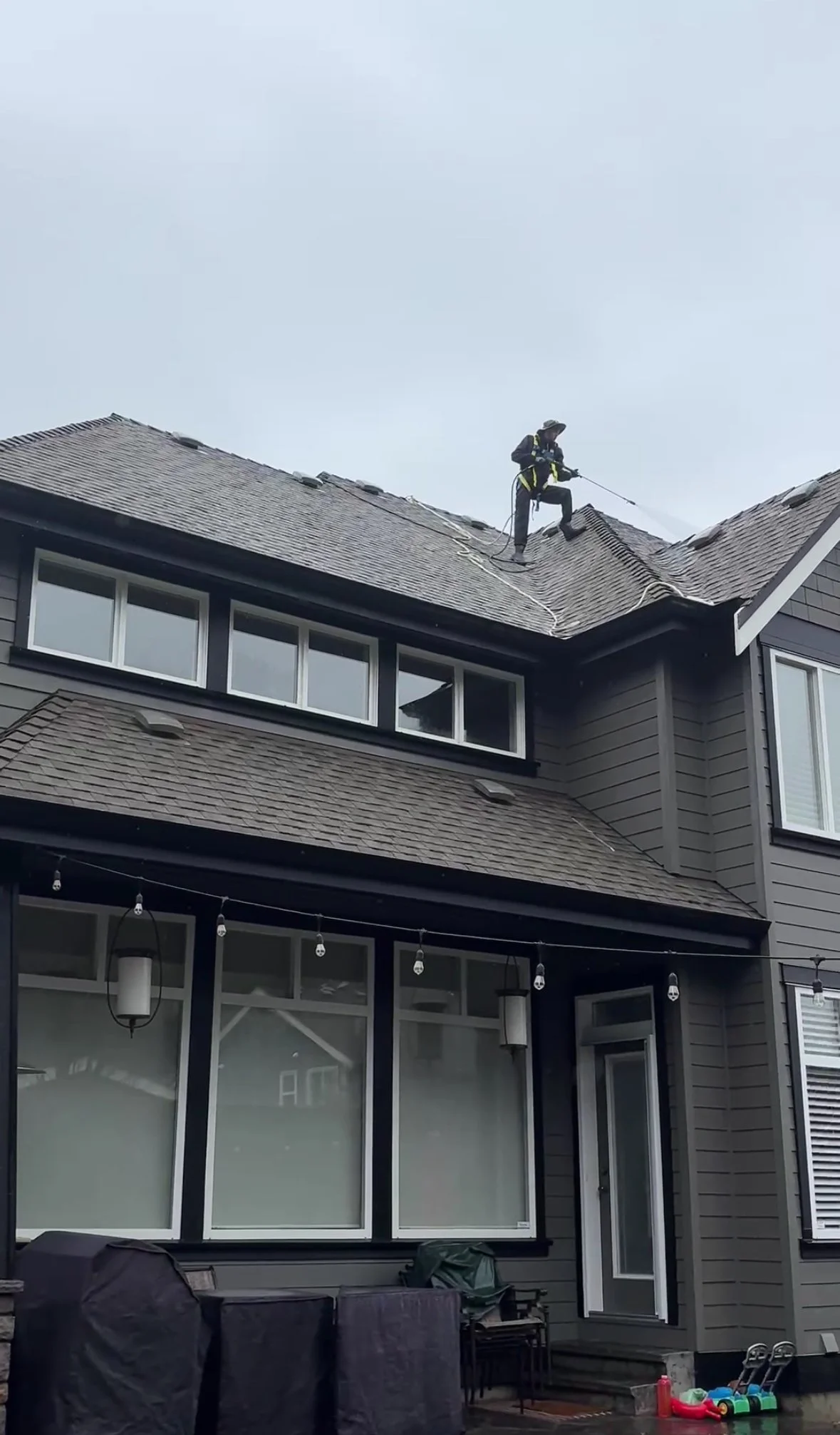 A person wearing safety gear cleaning or inspecting a residential house roof using a long pole or spray, standing near the ridge on a cloudy day.