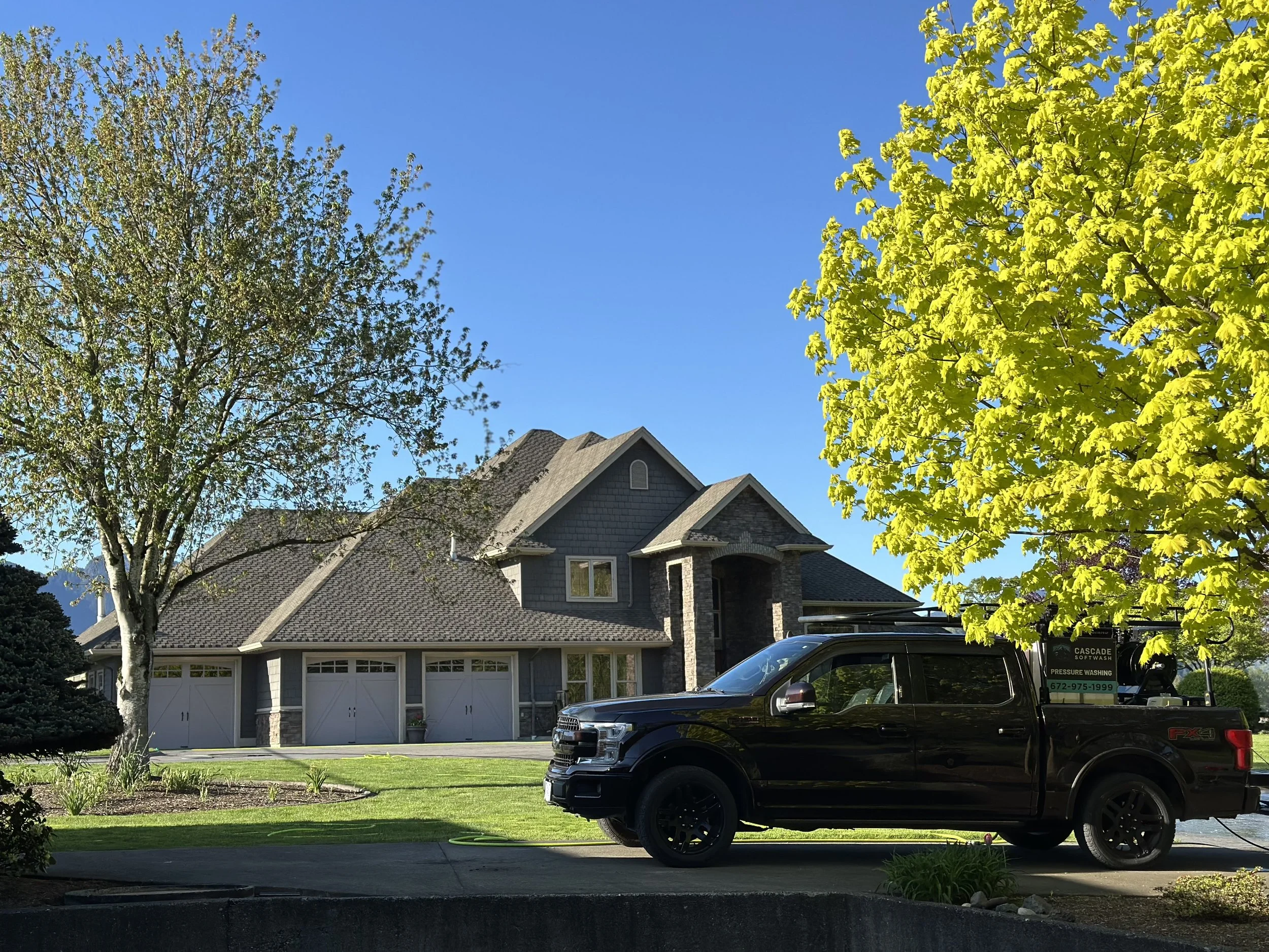 A large house with a gray shingle roof and white garage doors, surrounded by green lawn and trees, with a black pickup truck parked in front and a pressure washing sign on the vehicle.