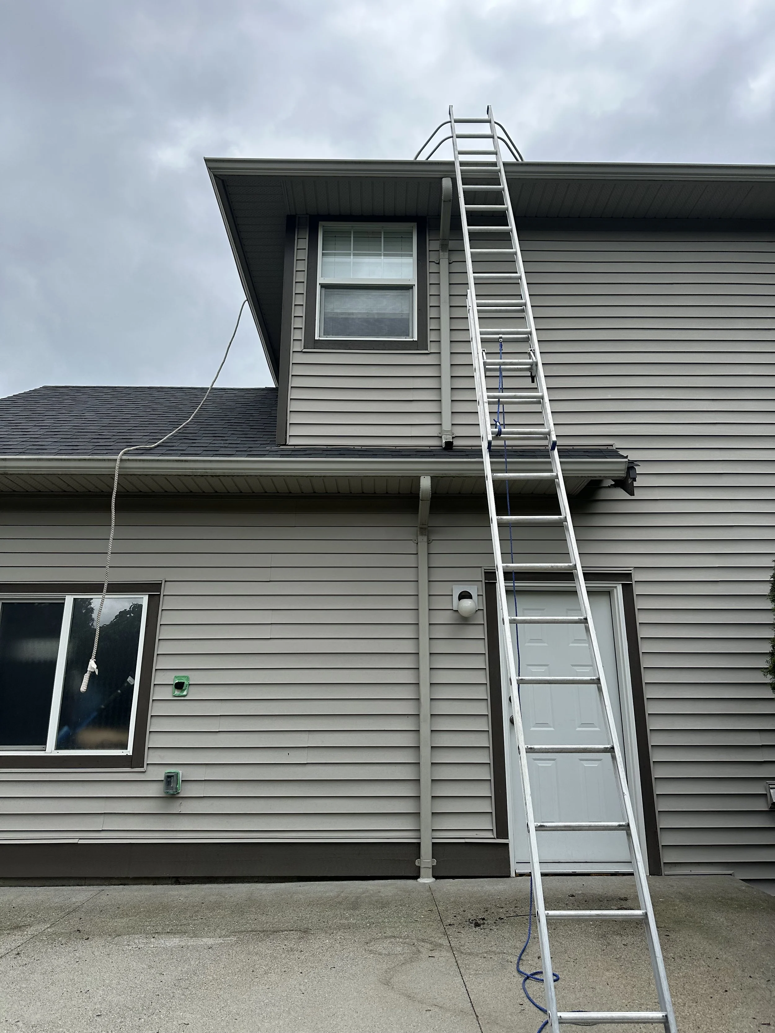 The side view of a house with two stories, beige siding, and a ladder leaning against the upper level. The house features a large blue-framed window on the second floor and a white door on the first floor. Cloudy sky in the background and concrete pa