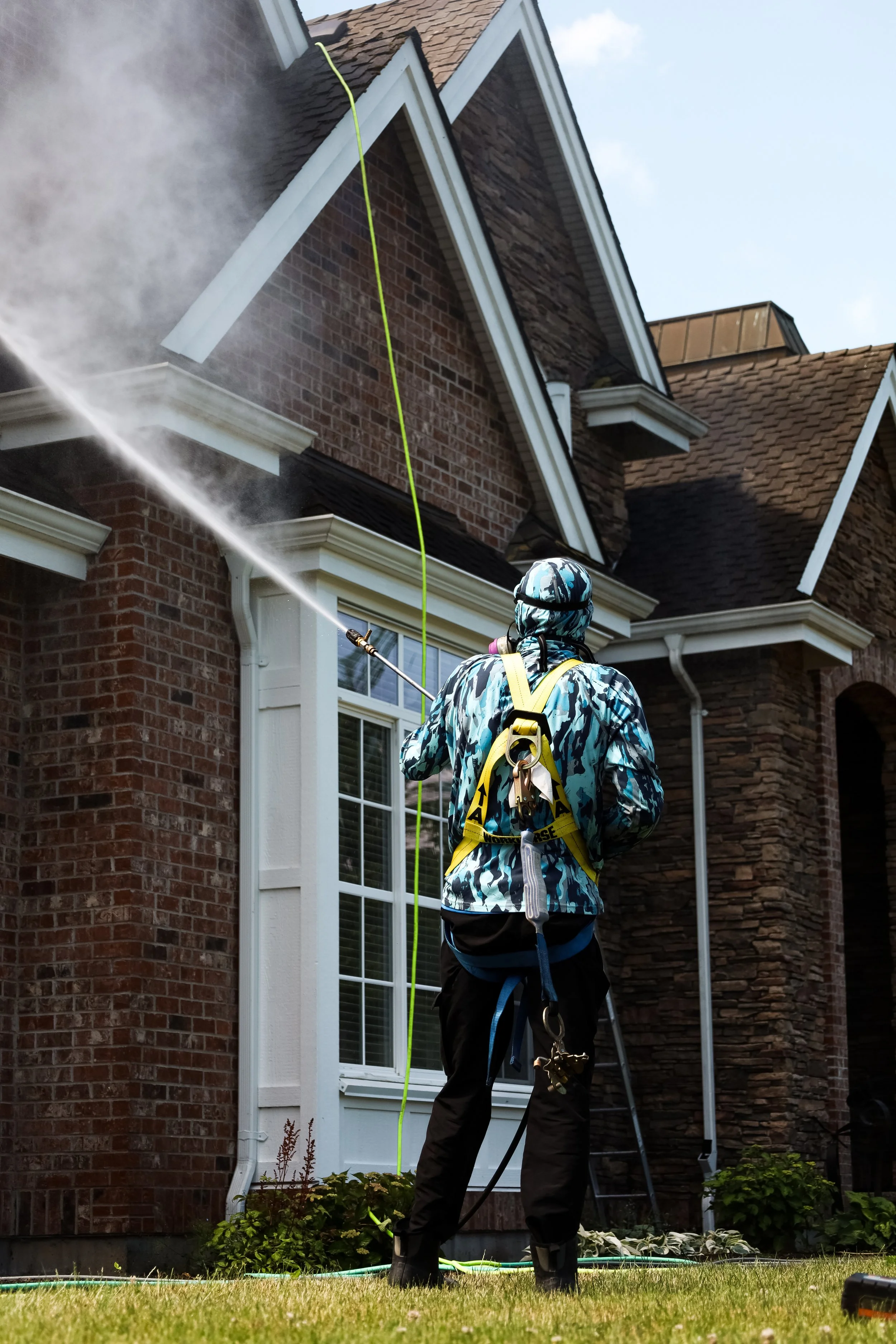 A person wearing a camouflage jacket and climbing harness is pressure washing the exterior of a brick house. The house has large windows with white frames, and the man is directing a high-pressure spray at the siding. The scene takes place in a well-
