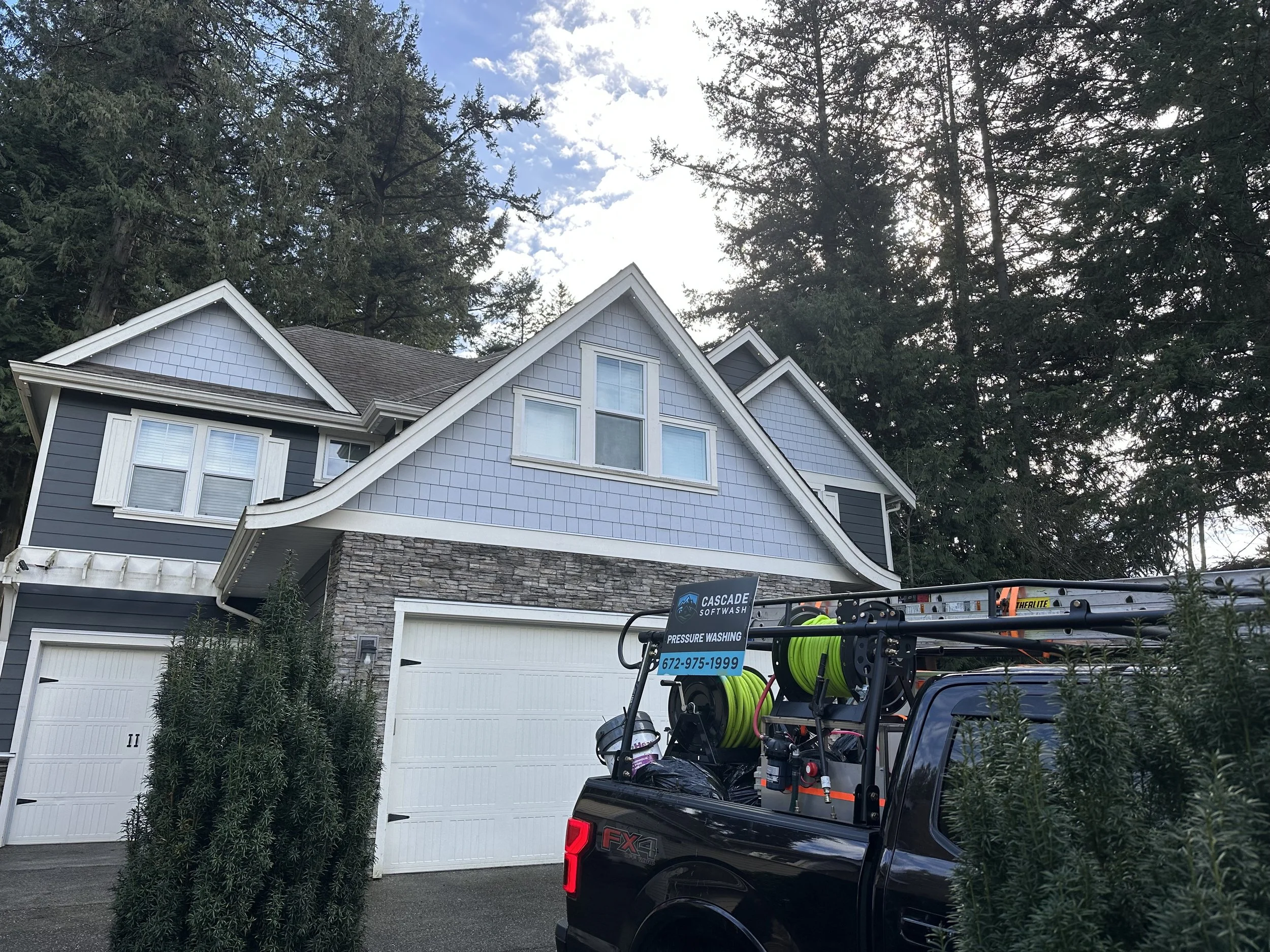A two-story house with light blue and dark blue siding, stone accents, three white garage doors, and a roof surrounded by tall trees. A black service truck with hose reels and equipment is parked in front.
