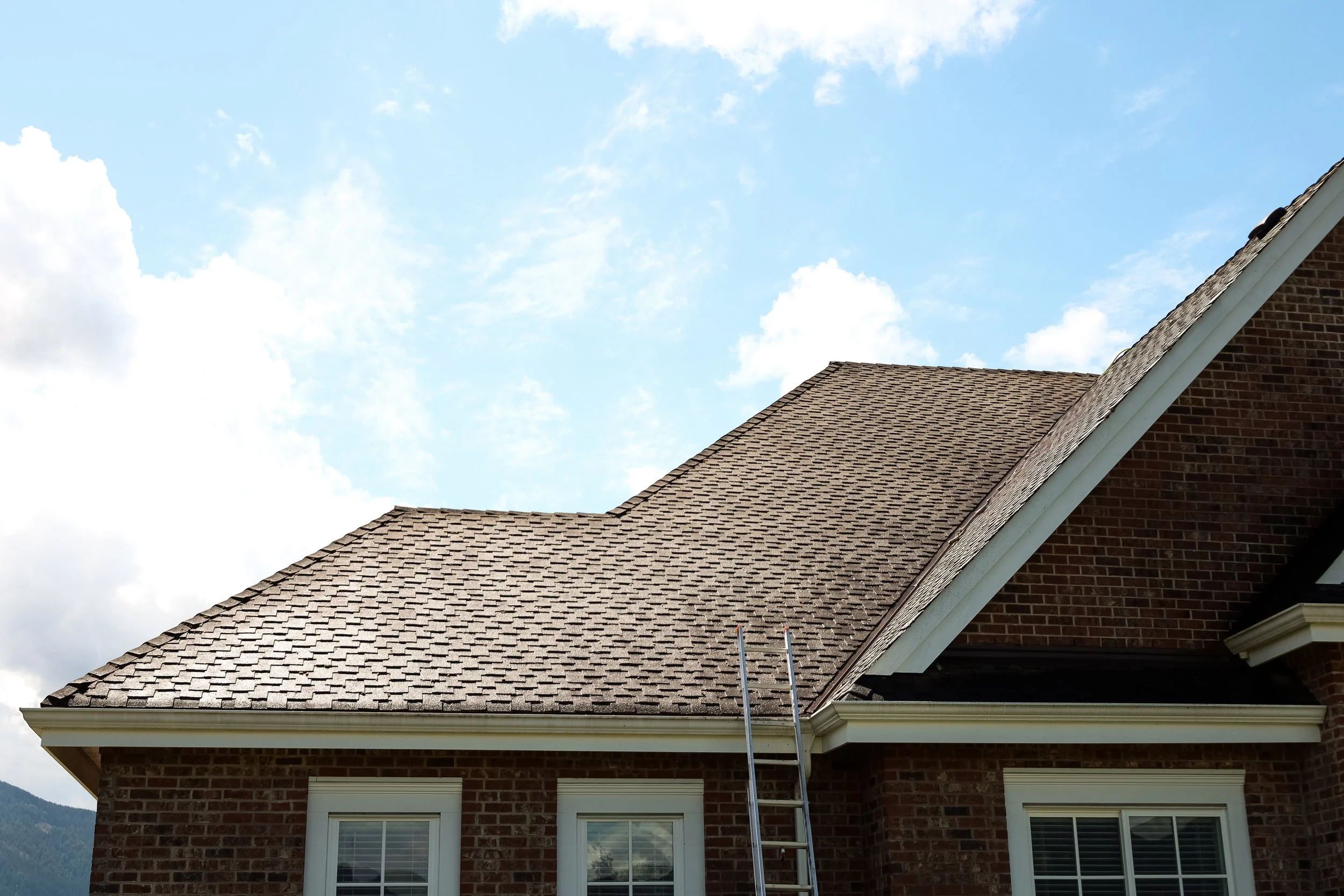 A residential house rooftop with brown shingles and brick walls, under a partly cloudy sky with white clouds.
