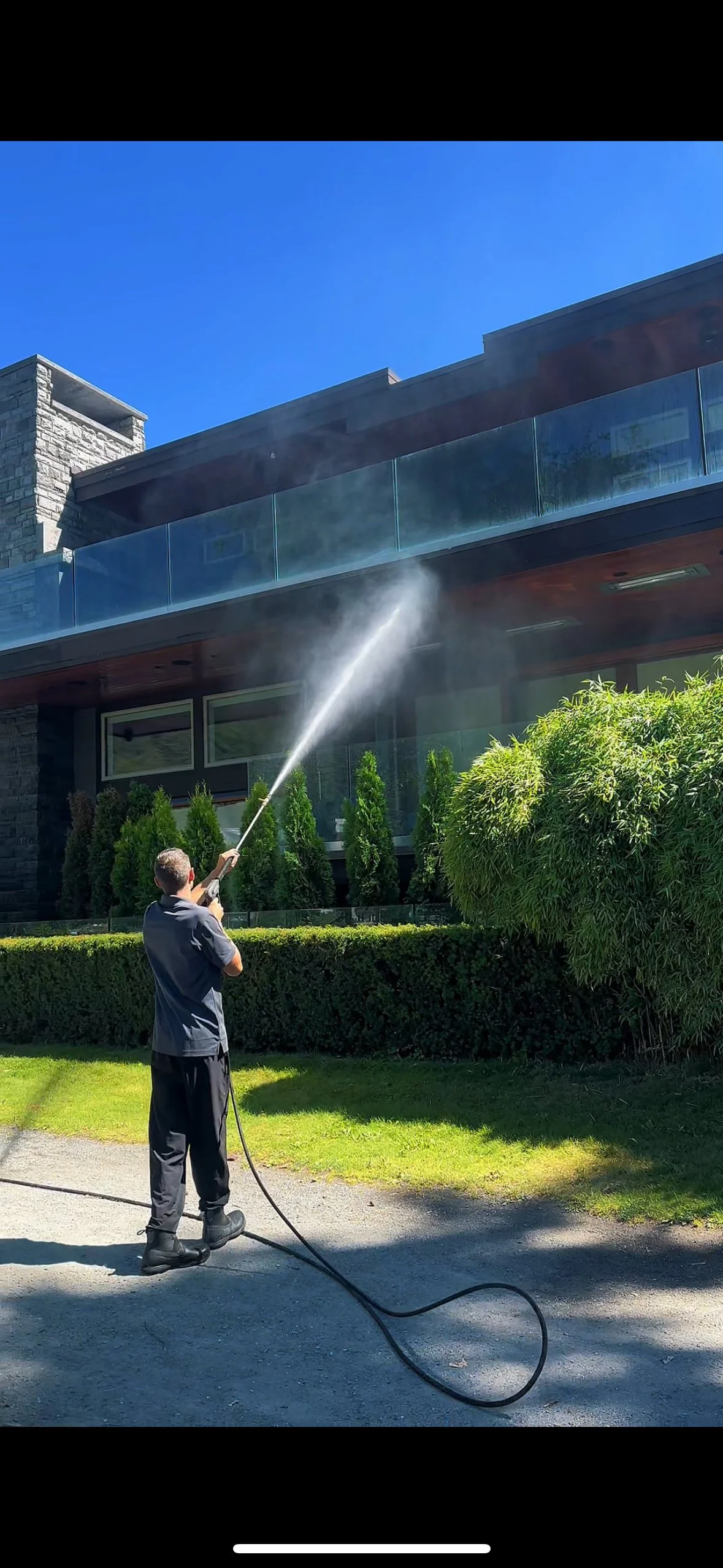 A man cleaning windows with a pressure washer on the exterior of a modern building with stone and glass architecture, green bushes, and clear blue sky.