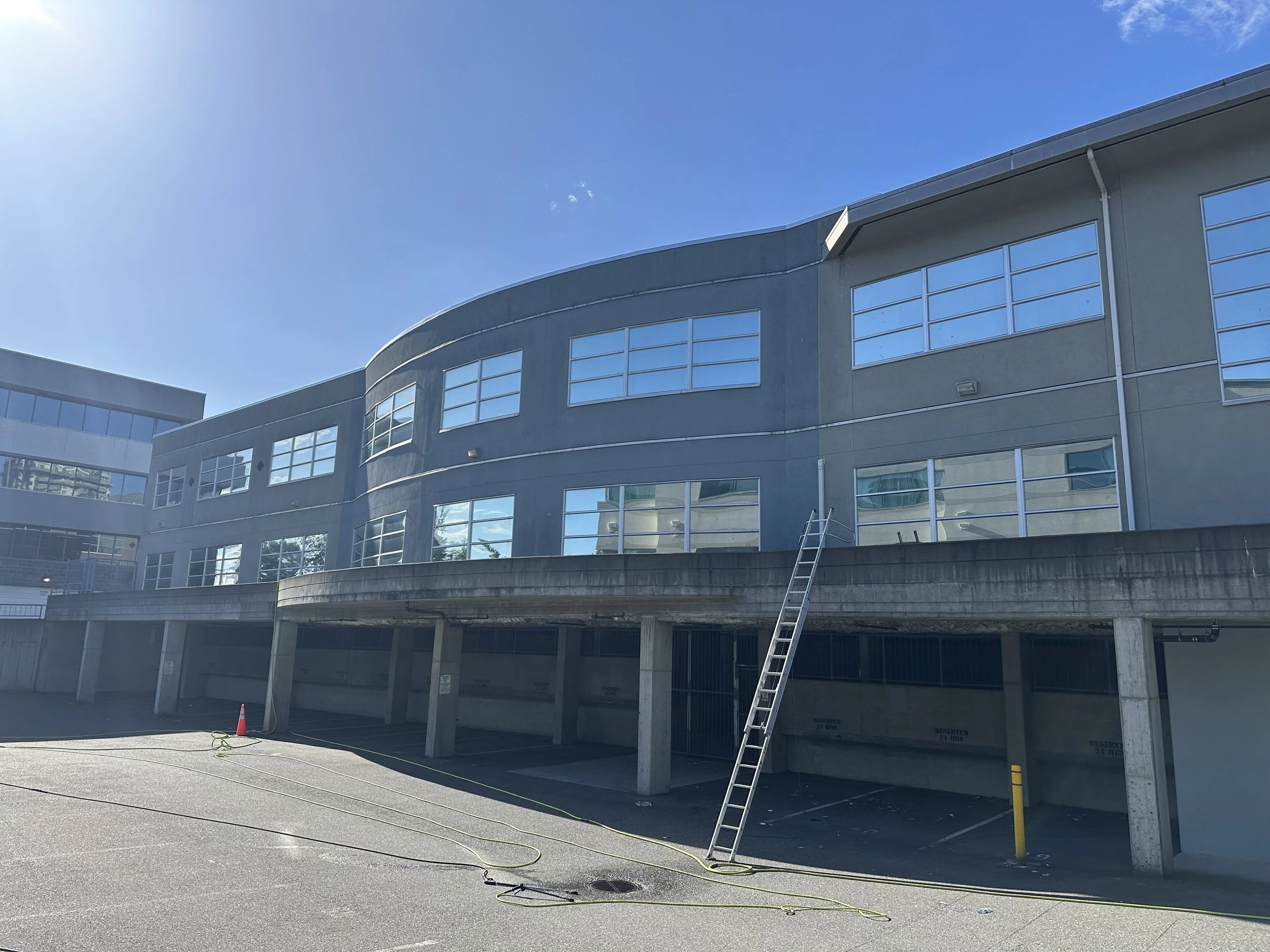 A modern multi-story building under construction with large glass windows and an exterior made of concrete or similar material. A ladder leans against the building, and construction cones and cables are present on the ground, with a parking lot in th